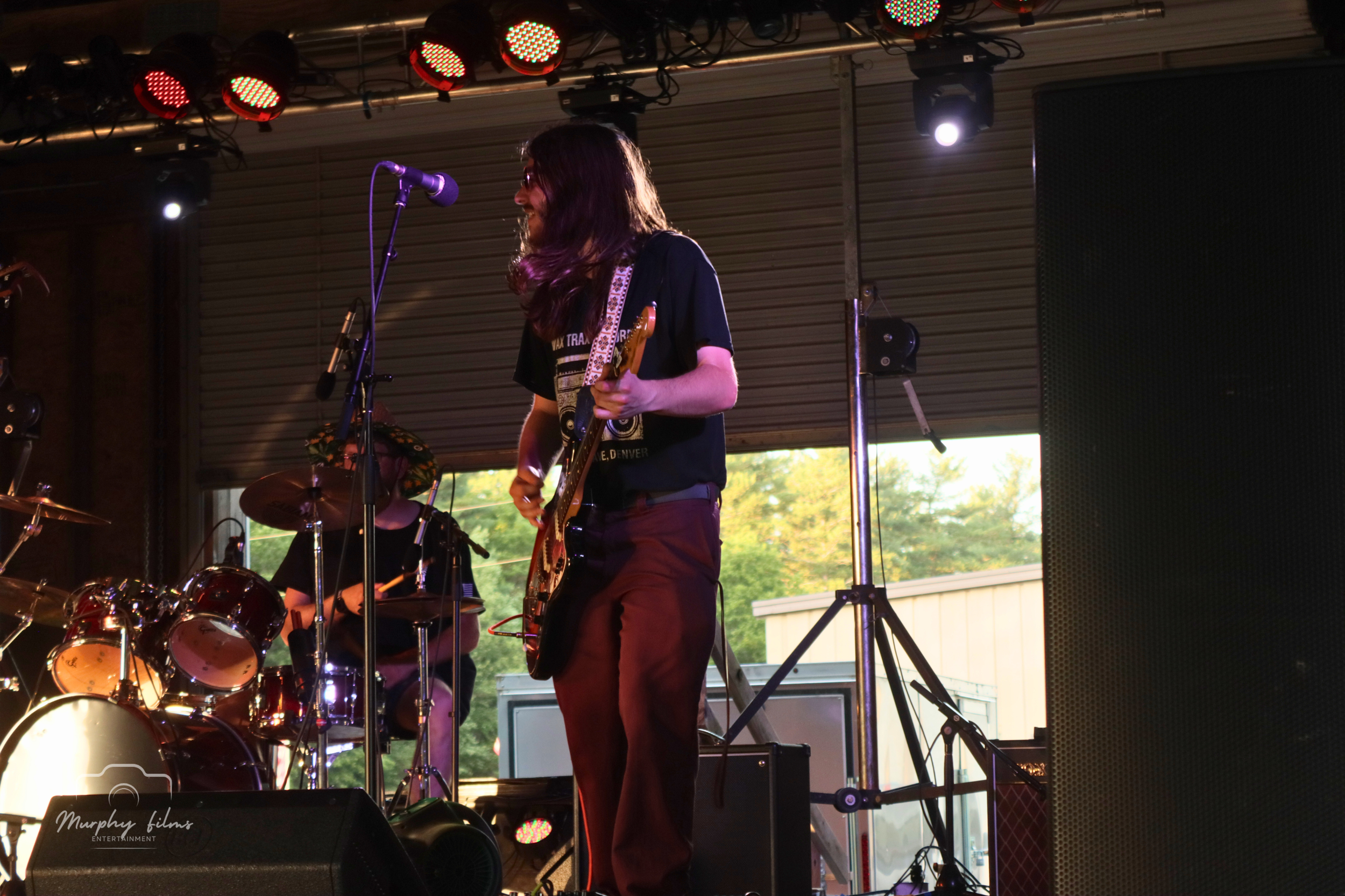 Musicians performing on stage, one playing guitar and another on drums, with stage lighting and equipment.