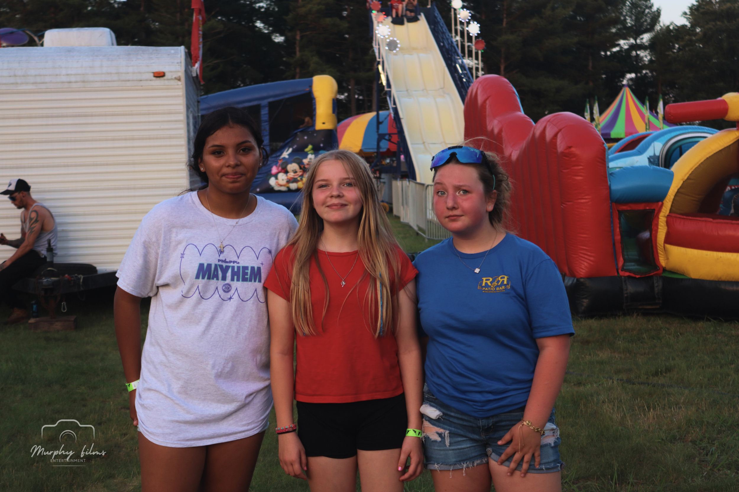 Three young girls standing together at an amusement park or fairground with inflatables and rides in the background.
