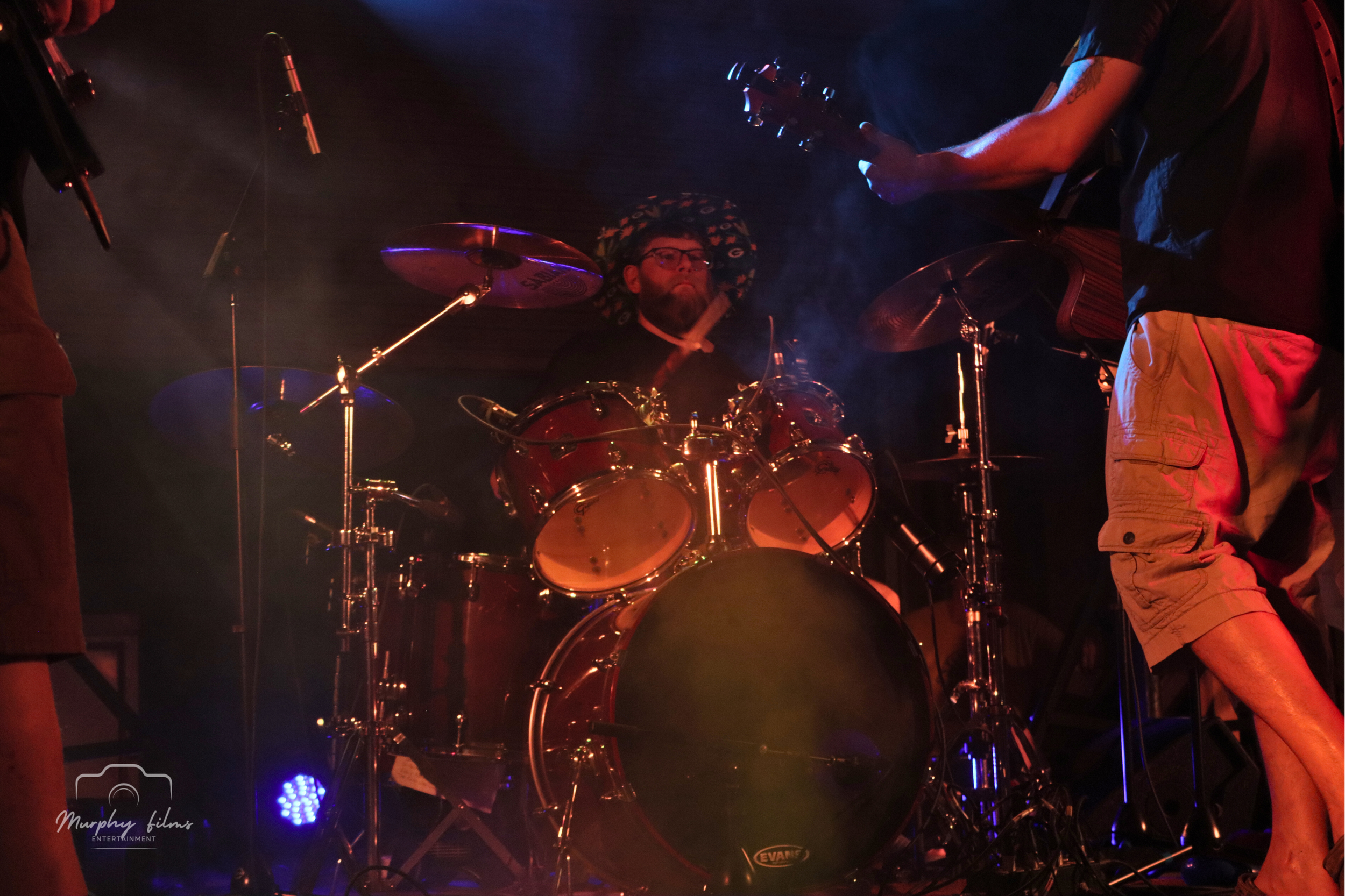 A person playing drums on stage with other musicians, wearing a hat with a pattern, glasses, and dark clothing, illuminated by stage lights.