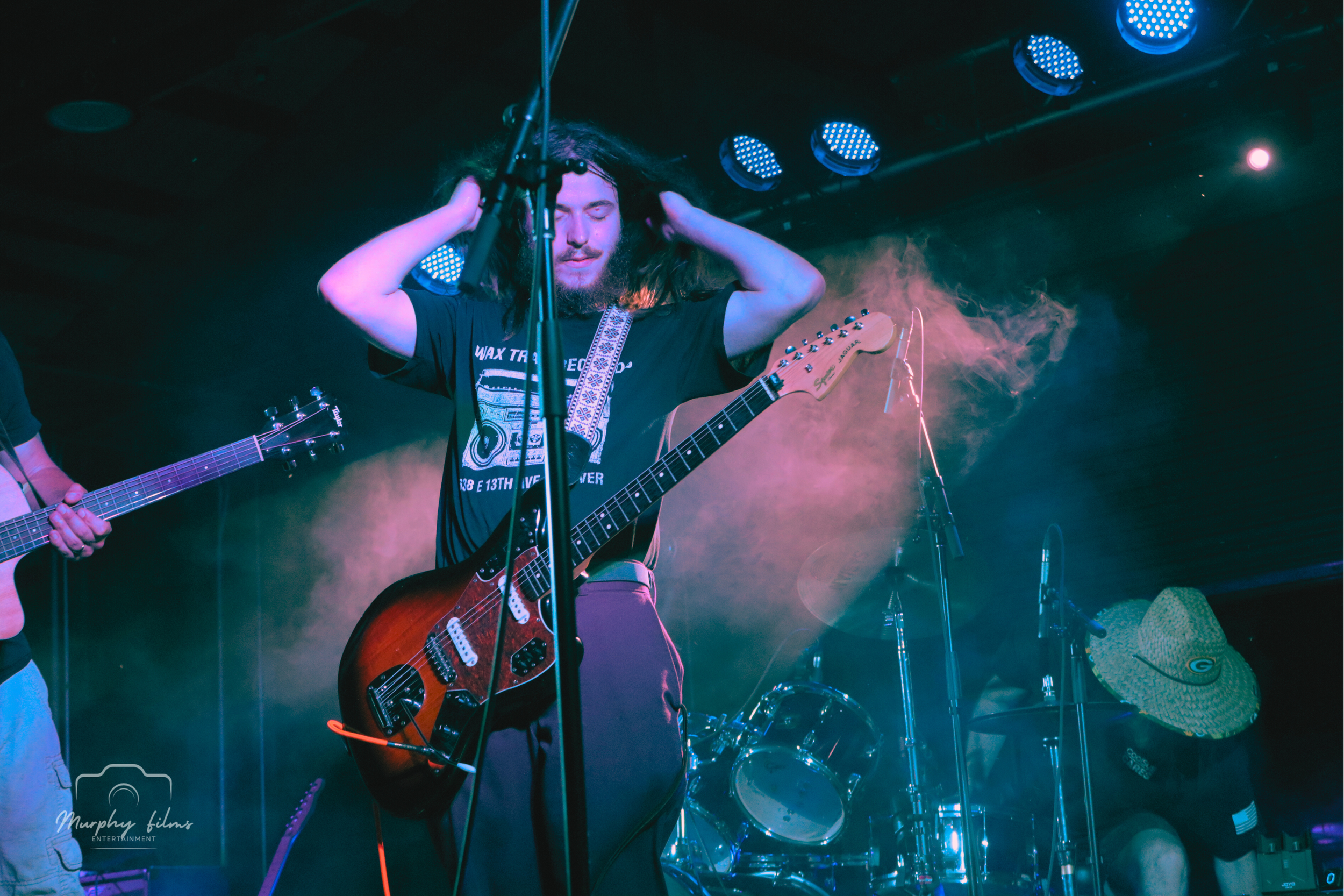 A musician with long dark hair, headphones on, eyes closed, playing an electric guitar on stage with colorful lights and smoke, while another person wearing a straw hat and backpack is adjusting equipment nearby.