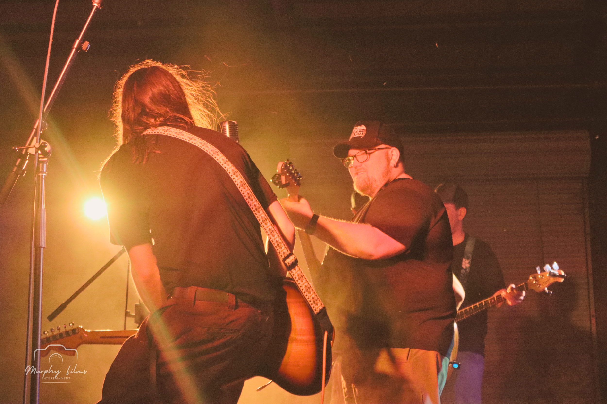 Two men on stage playing guitars, one with long hair and the other with a cap and glasses, with warm stage lighting creating a cozy atmosphere.