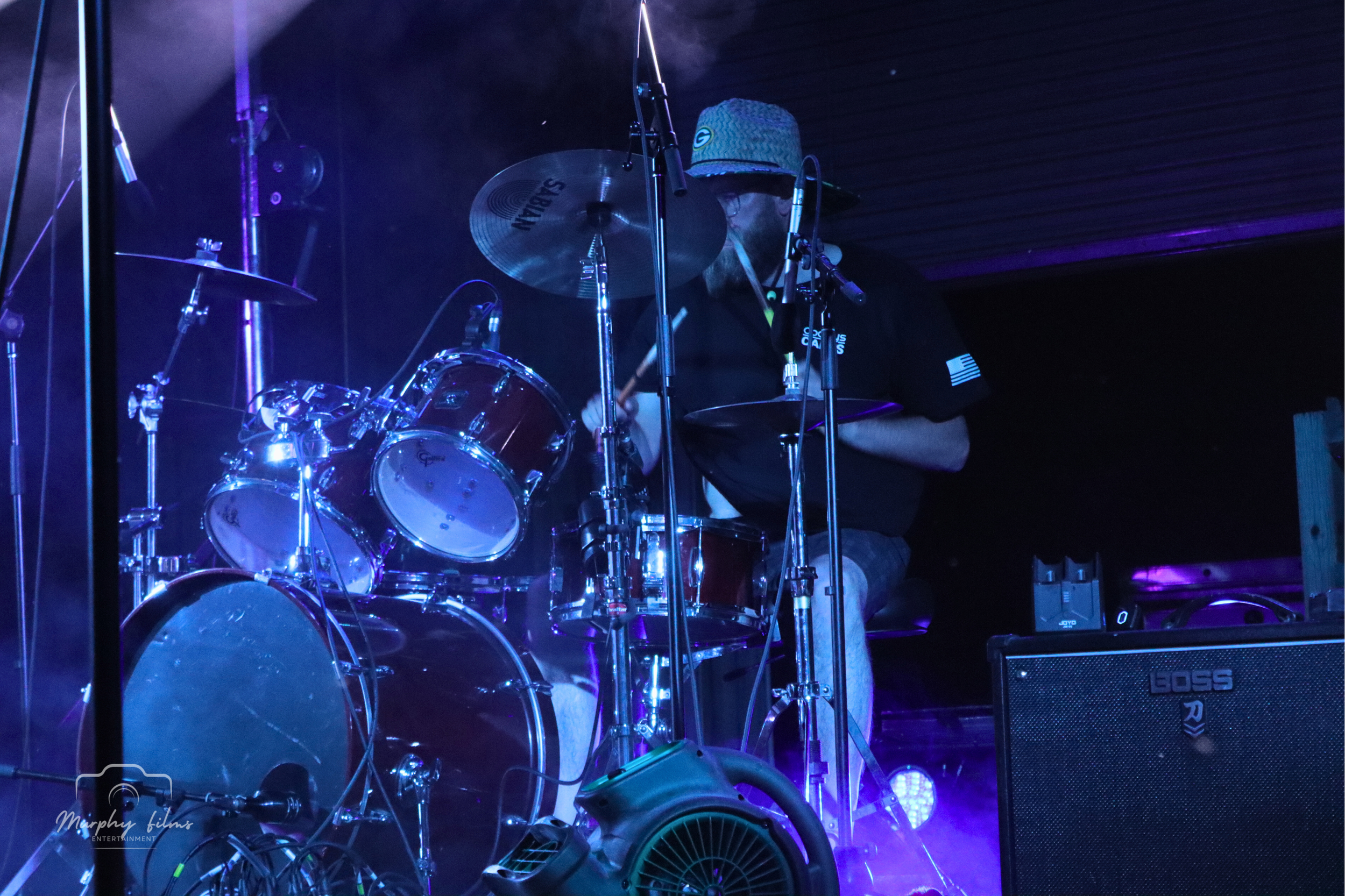 A drummer playing a drum kit on stage illuminated by purple and blue lights, with a BOSS amplifier visible.