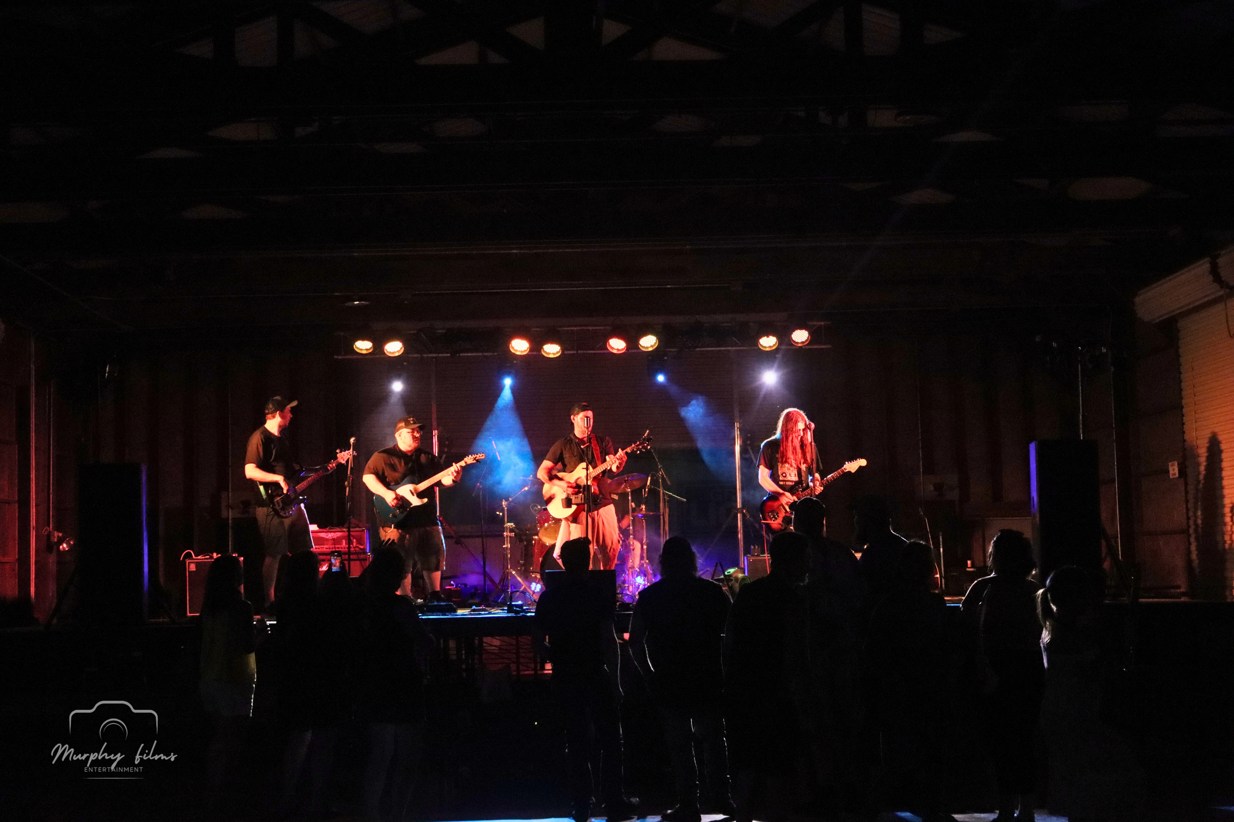 A band performing on a dimly lit stage with five musicians playing electric guitars, and a drummer in the background, illuminated by stage lights. Audience silhouettes are visible in the foreground.