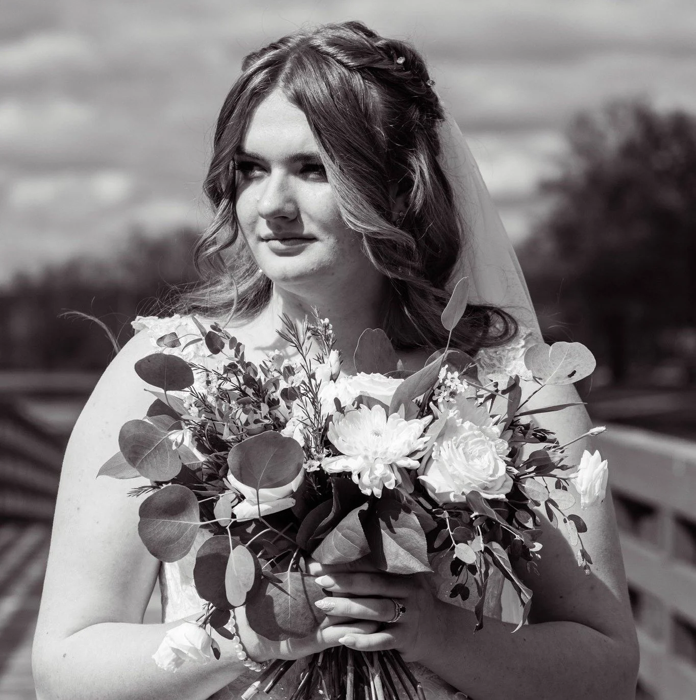 Black and white photo of a bride holding a bouquet of flowers, looking off to the side outdoors.
