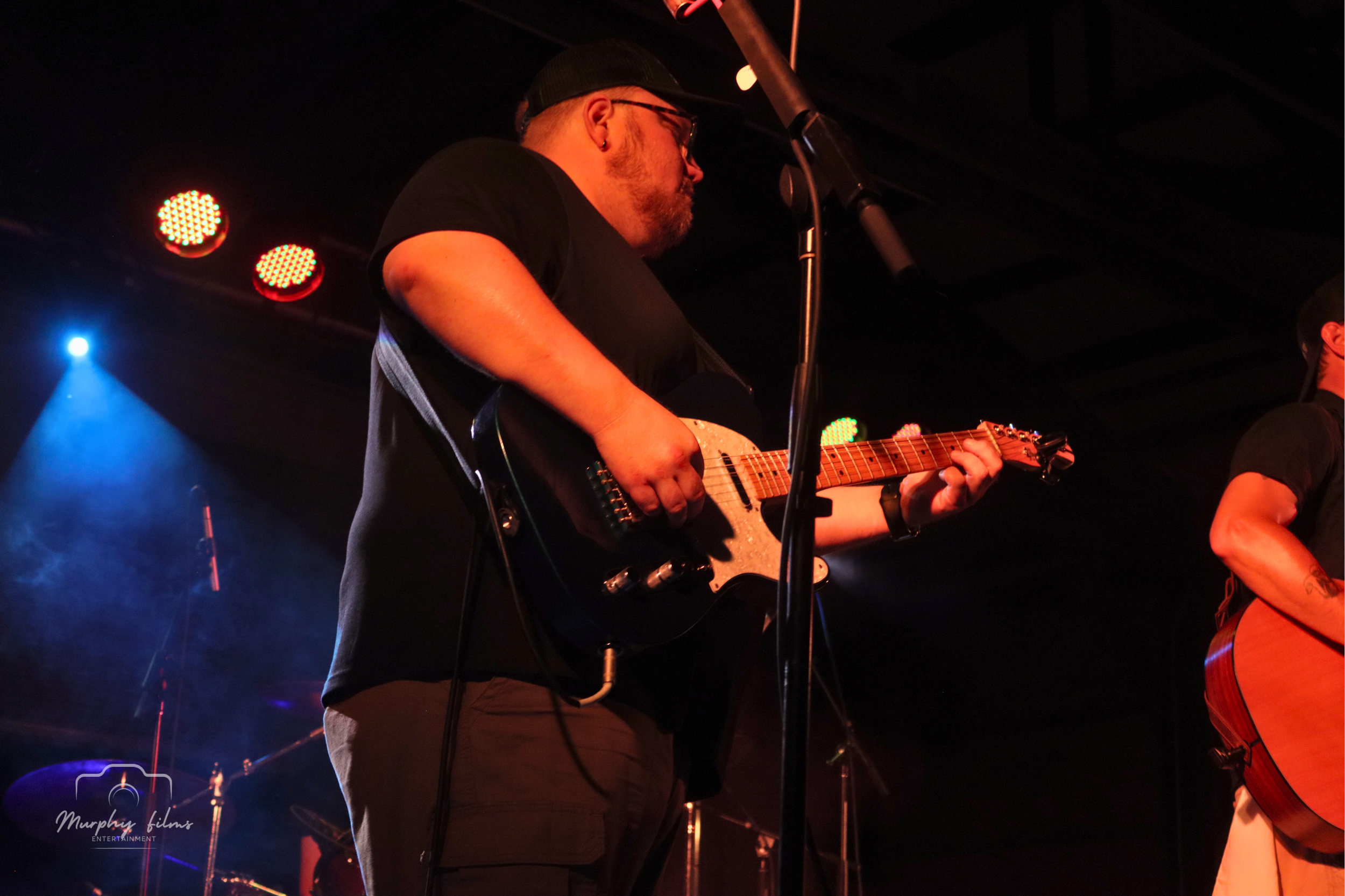 A male musician playing an electric guitar on stage, illuminated by stage lights, with a drum set and colorful lights in the background.