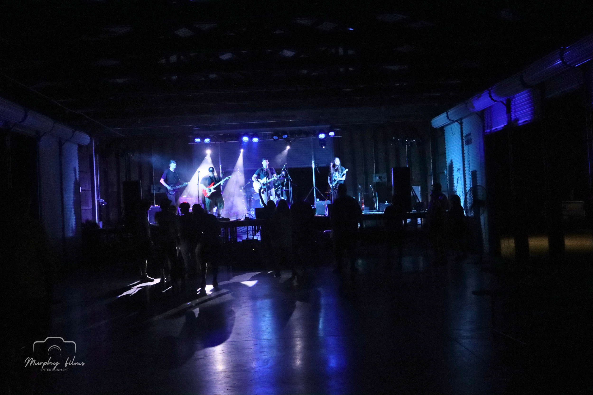 A band performing on stage with four guitarists and drums, illuminated by blue and purple stage lights, with silhouettes of an audience watching the performance.