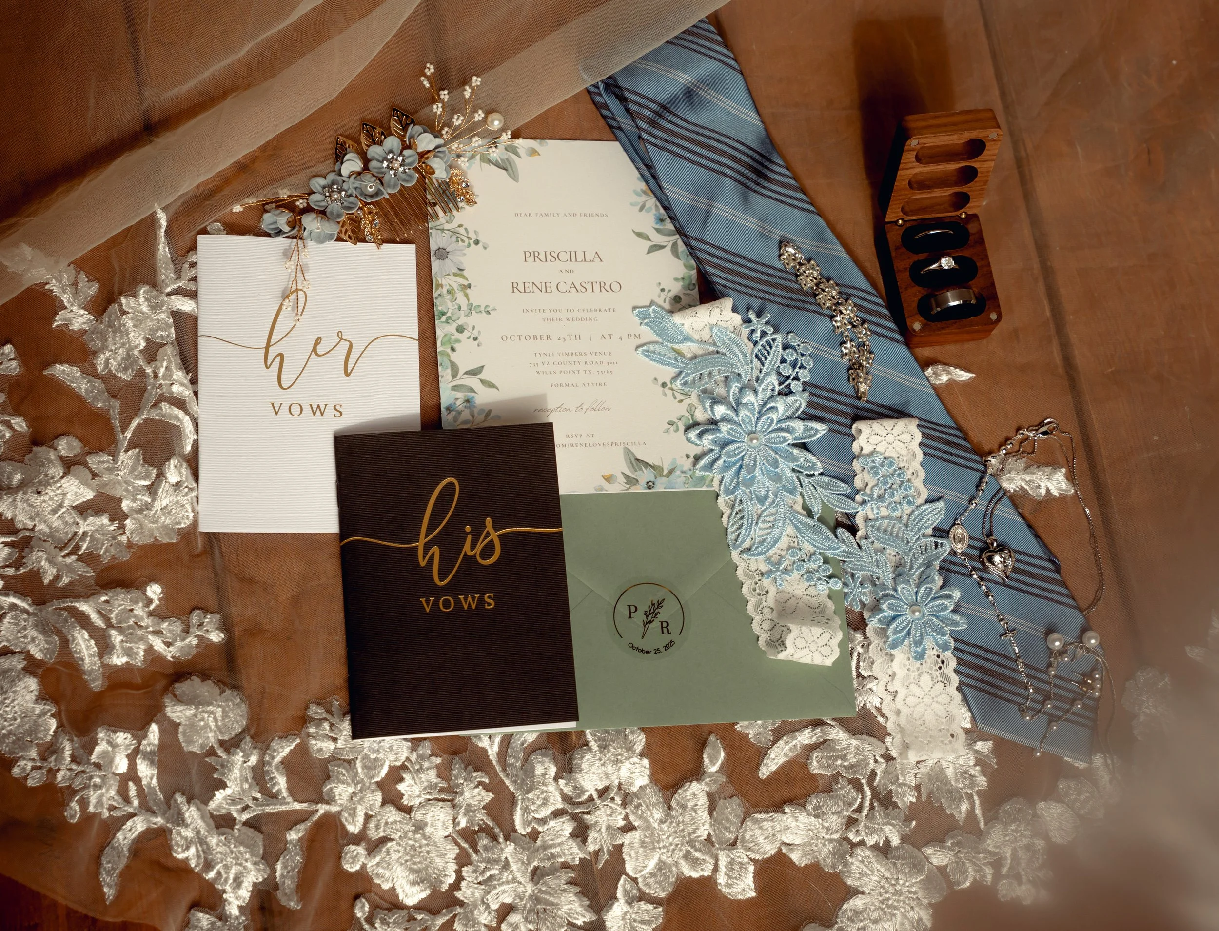 Wedding invitation suite with vows booklets, floral accessories, jewelry, and lace on a wooden table covered with a lace table runner.