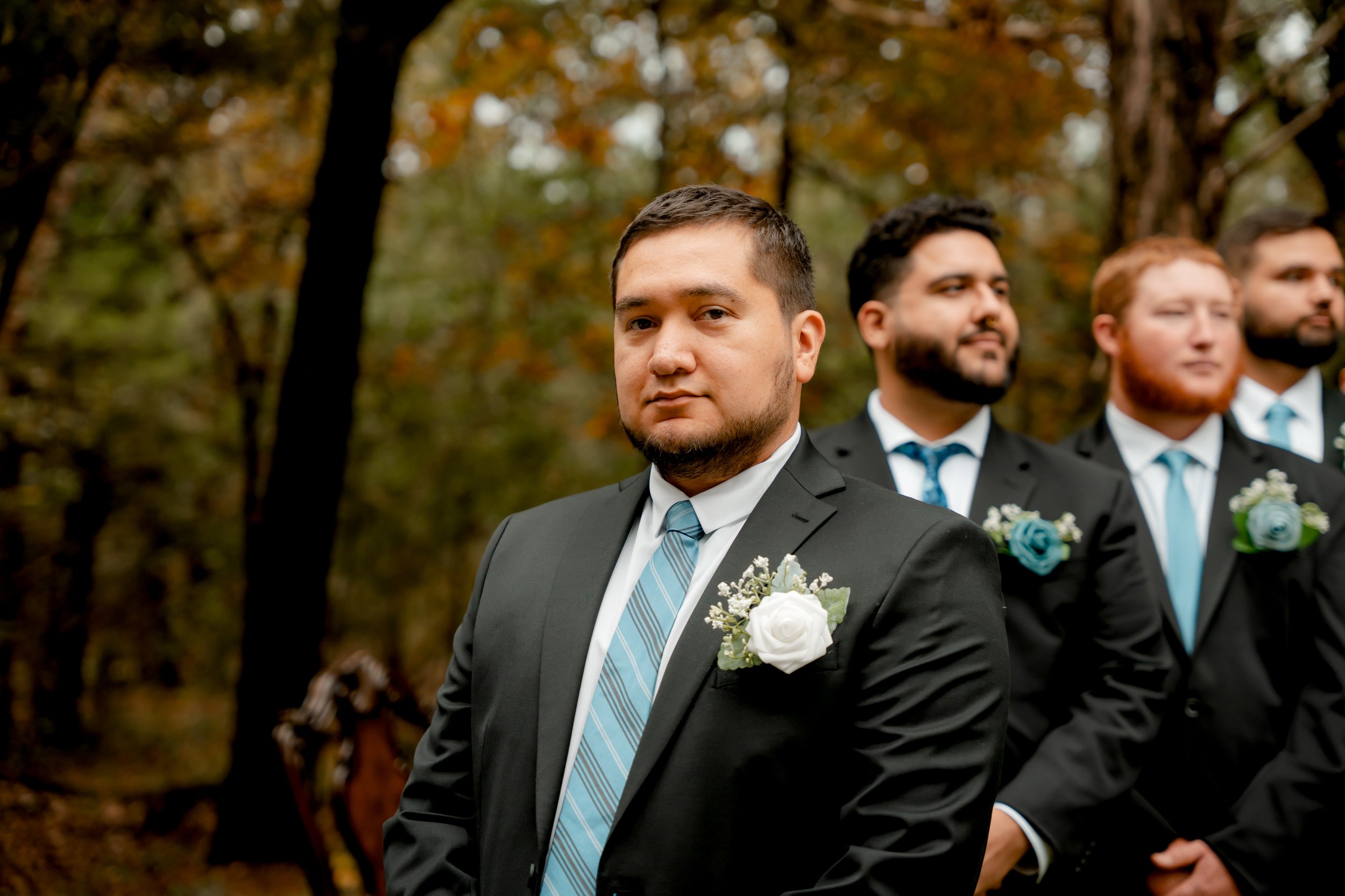 Group of men in suits with boutonnières, standing outdoors in a forested area during fall.