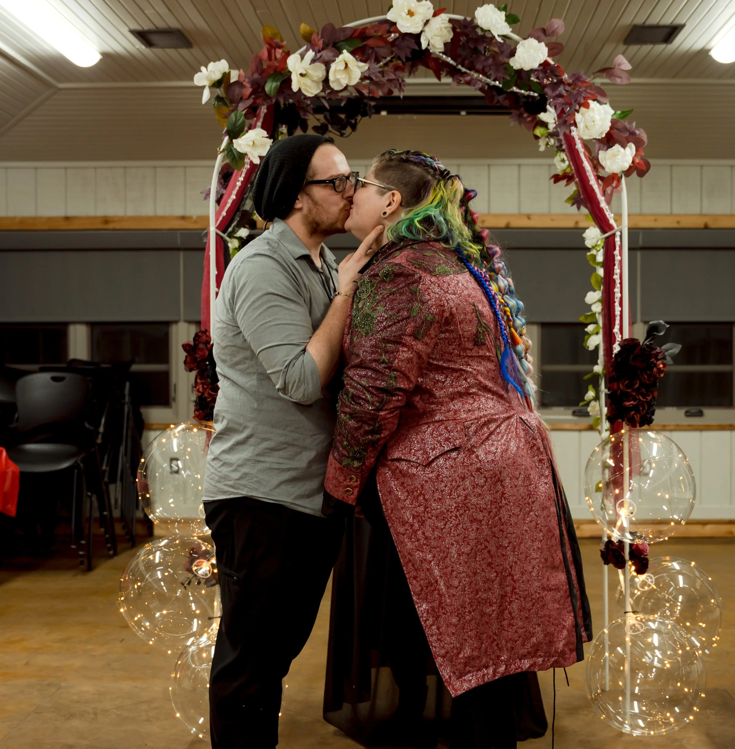 Two people kissing under a floral arch at a celebration or wedding, with balloons and fairy lights in the background.