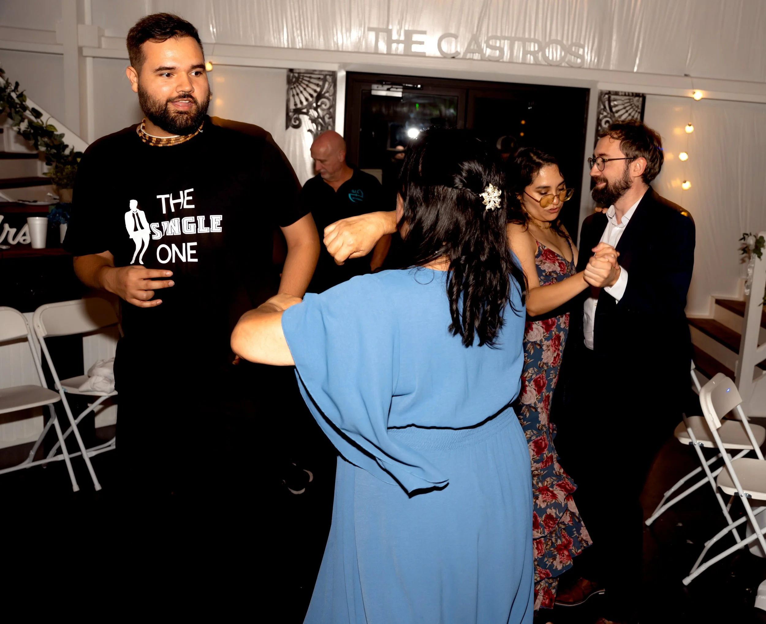 People dancing at a social gathering in a decorated indoor venue, with some sitting on white chairs and others dancing.