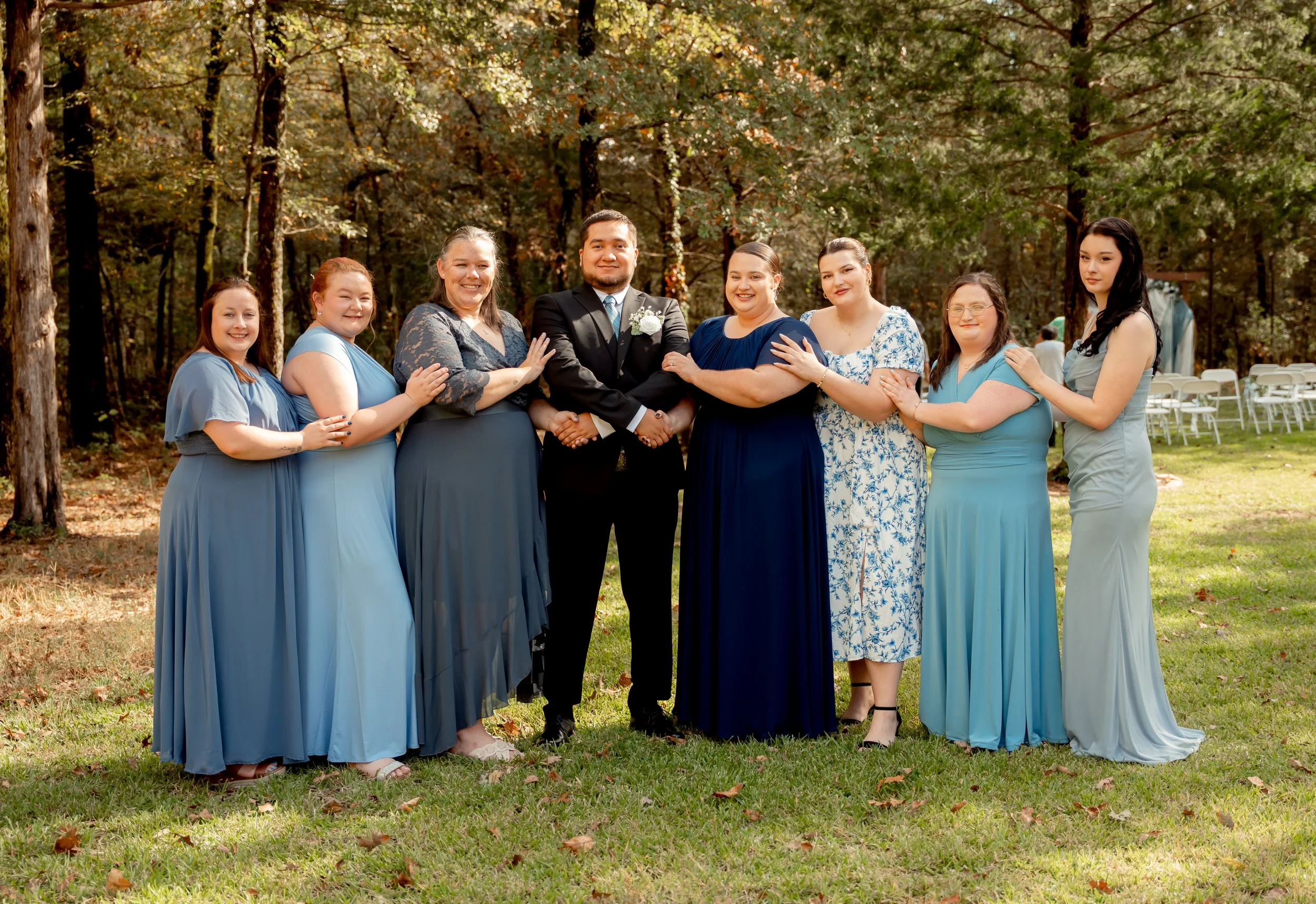 A group of women in blue dresses and a man in a black suit posing outdoors in a wooded area during daytime.