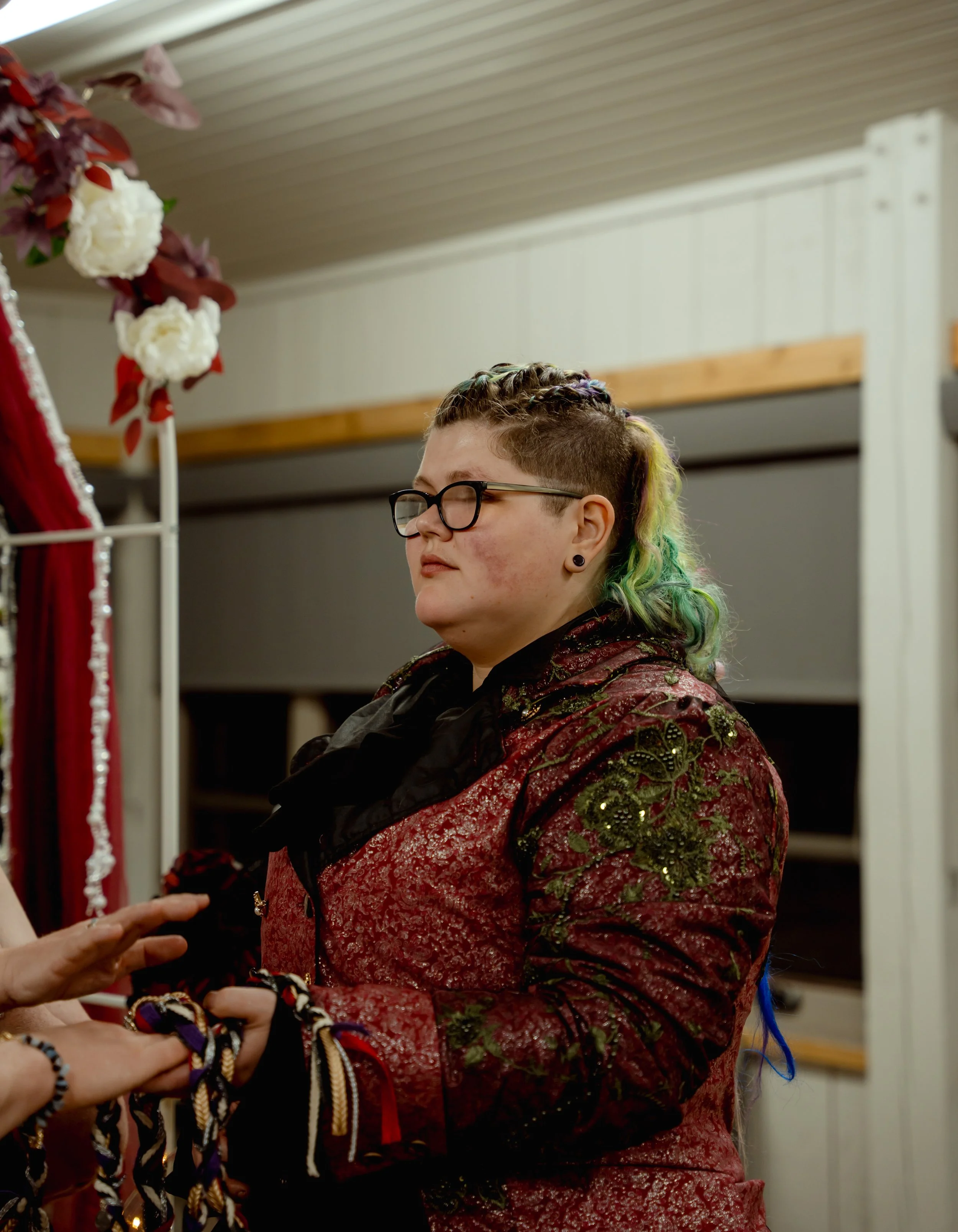A person with multicolored hair and glasses wearing an ornate red and green patterned jacket, standing indoors near a floral decoration.