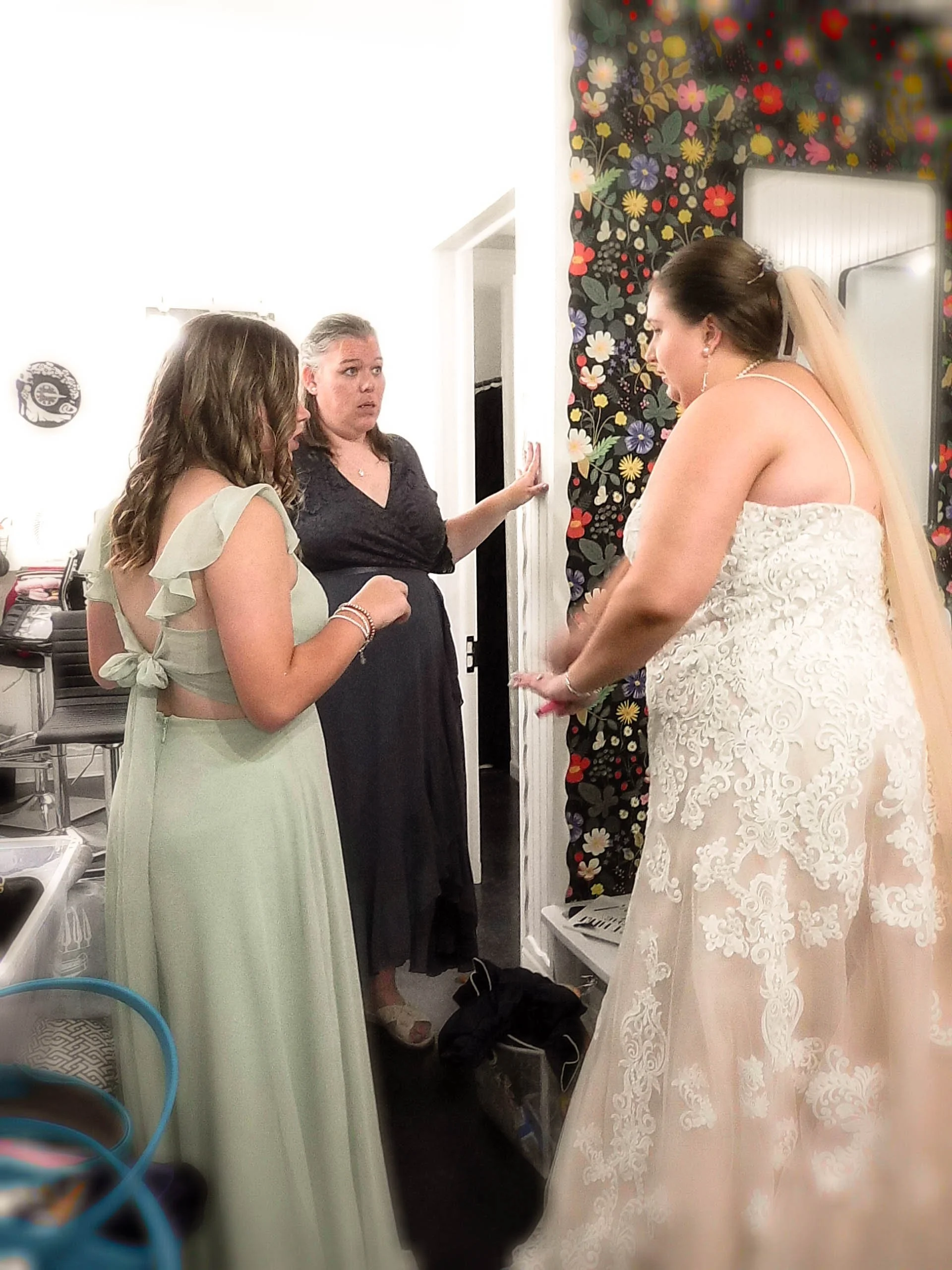 Three women in a room, two in casual dresses and one in a wedding dress, engaging in conversation with expressions of concern or surprise.