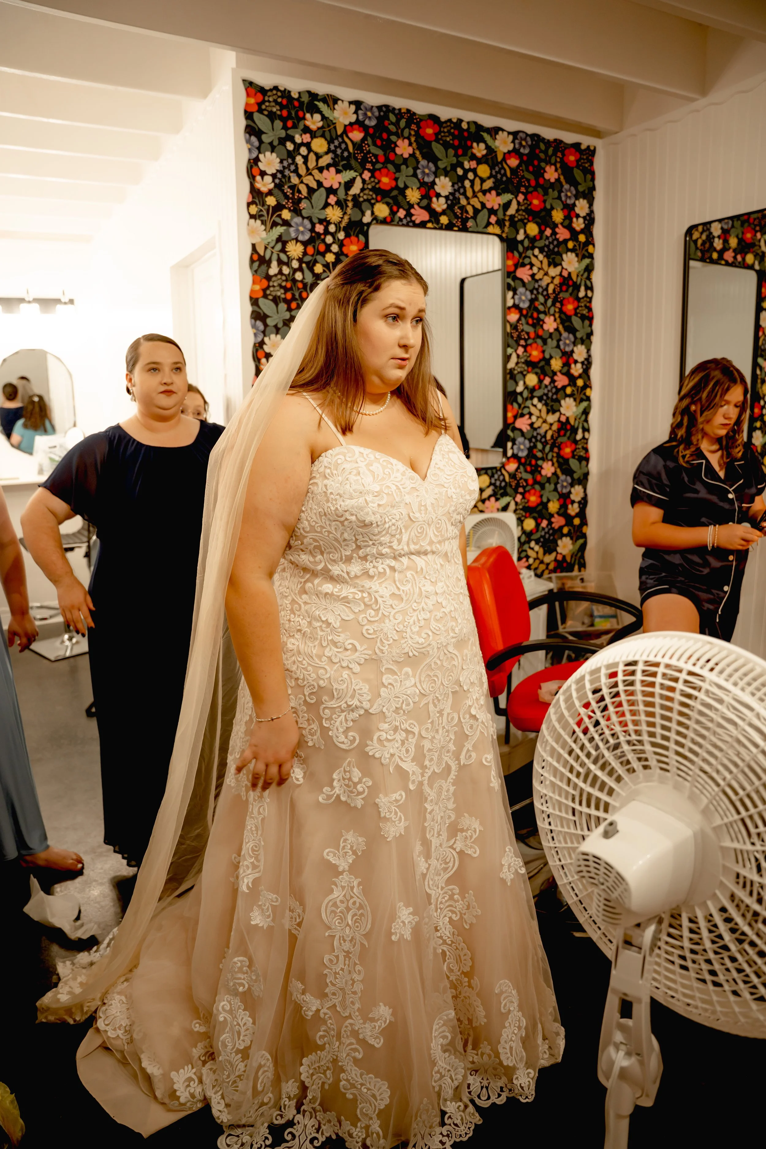 Bridal woman in a white lace wedding dress and veil standing in a room with other women, preparing for her wedding.