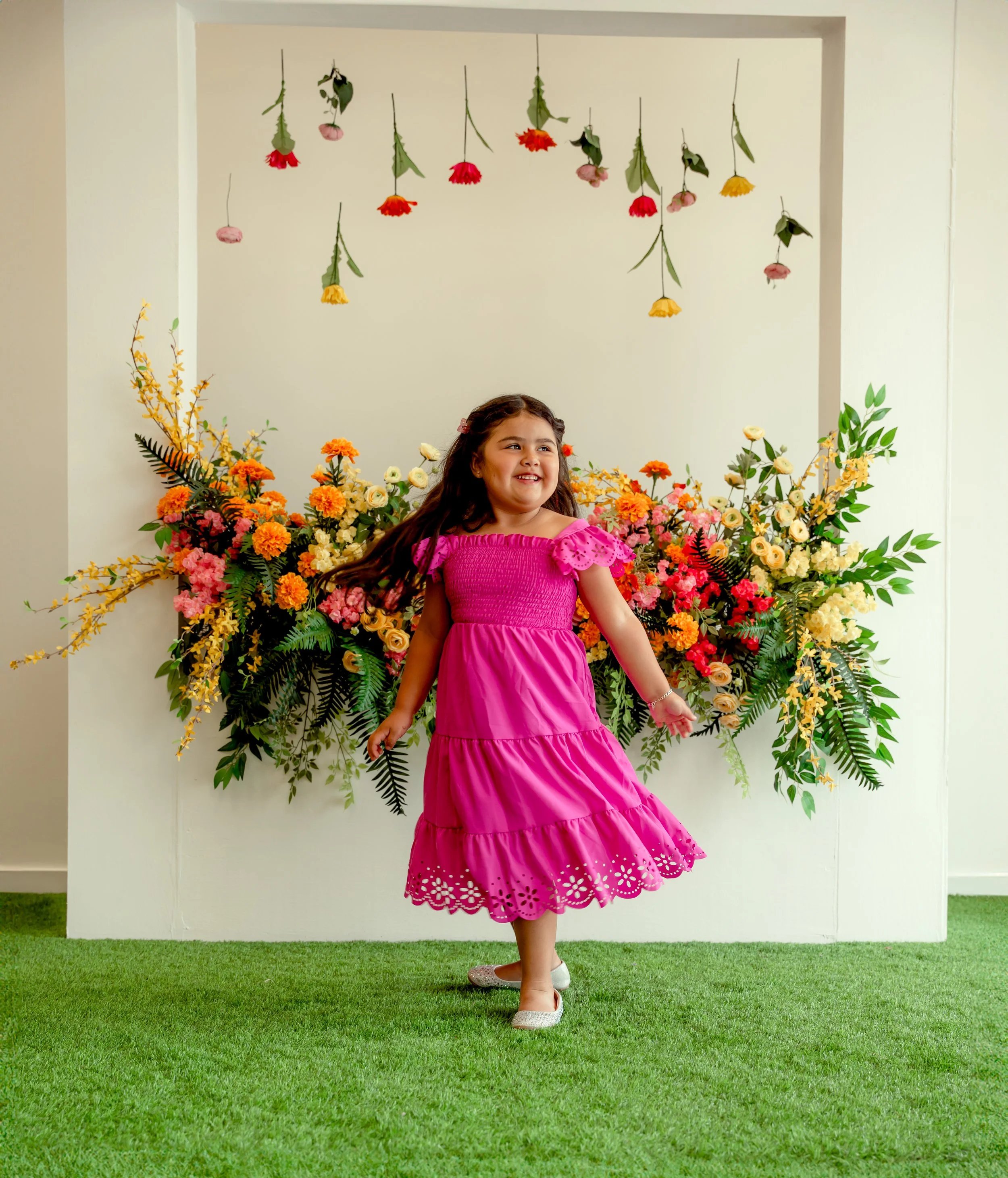 A young girl in a pink dress standing on green grass in front of a large floral arrangement, with hanging flowers above her and a white wall background.