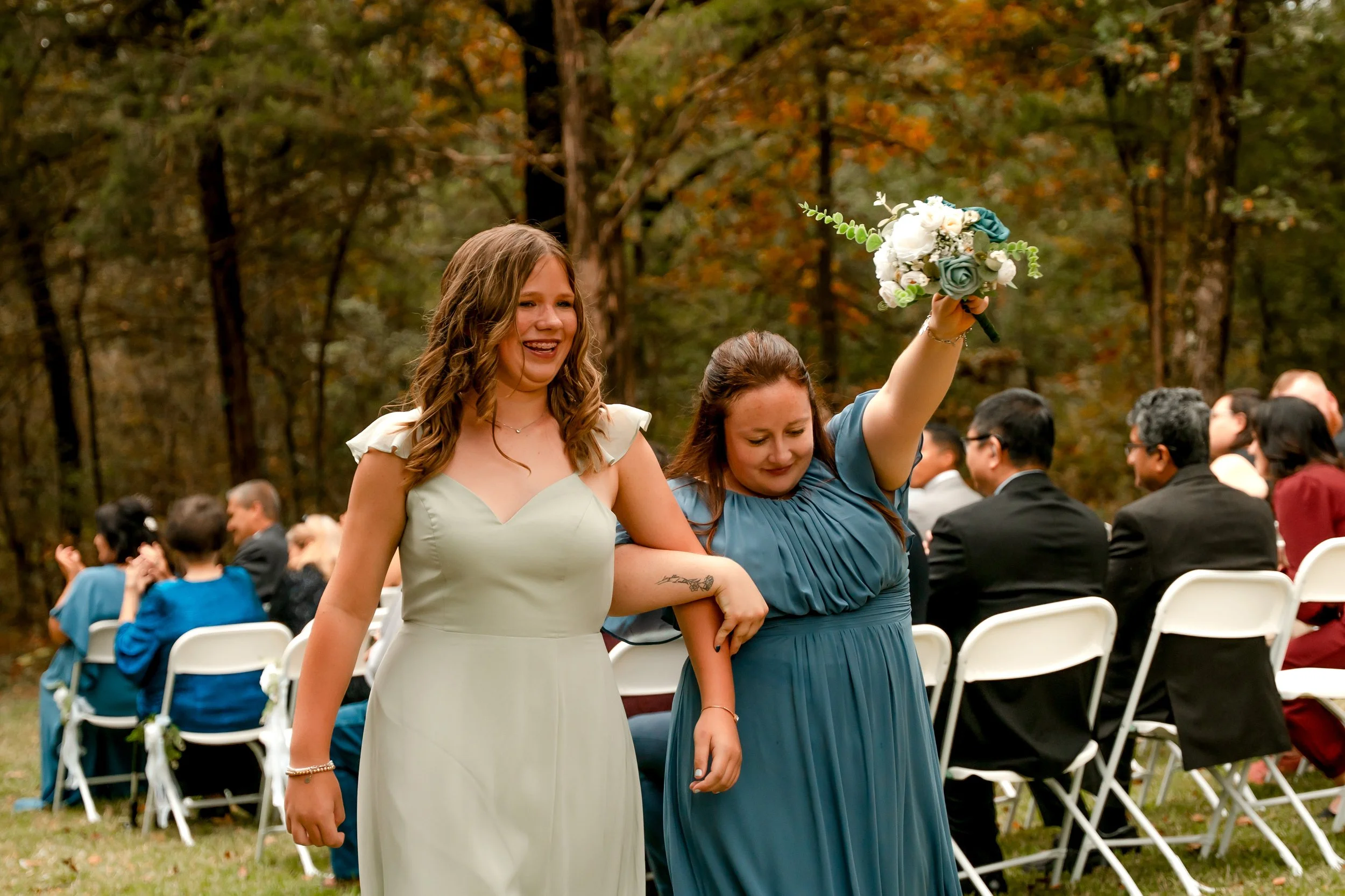 Two women, one in a pale green dress and the other in a teal dress, hold hands as they walk through an outdoor wedding ceremony. The woman in the teal dress holds a bouquet of white, teal, and green flowers and appears emotional. Guests are seated on