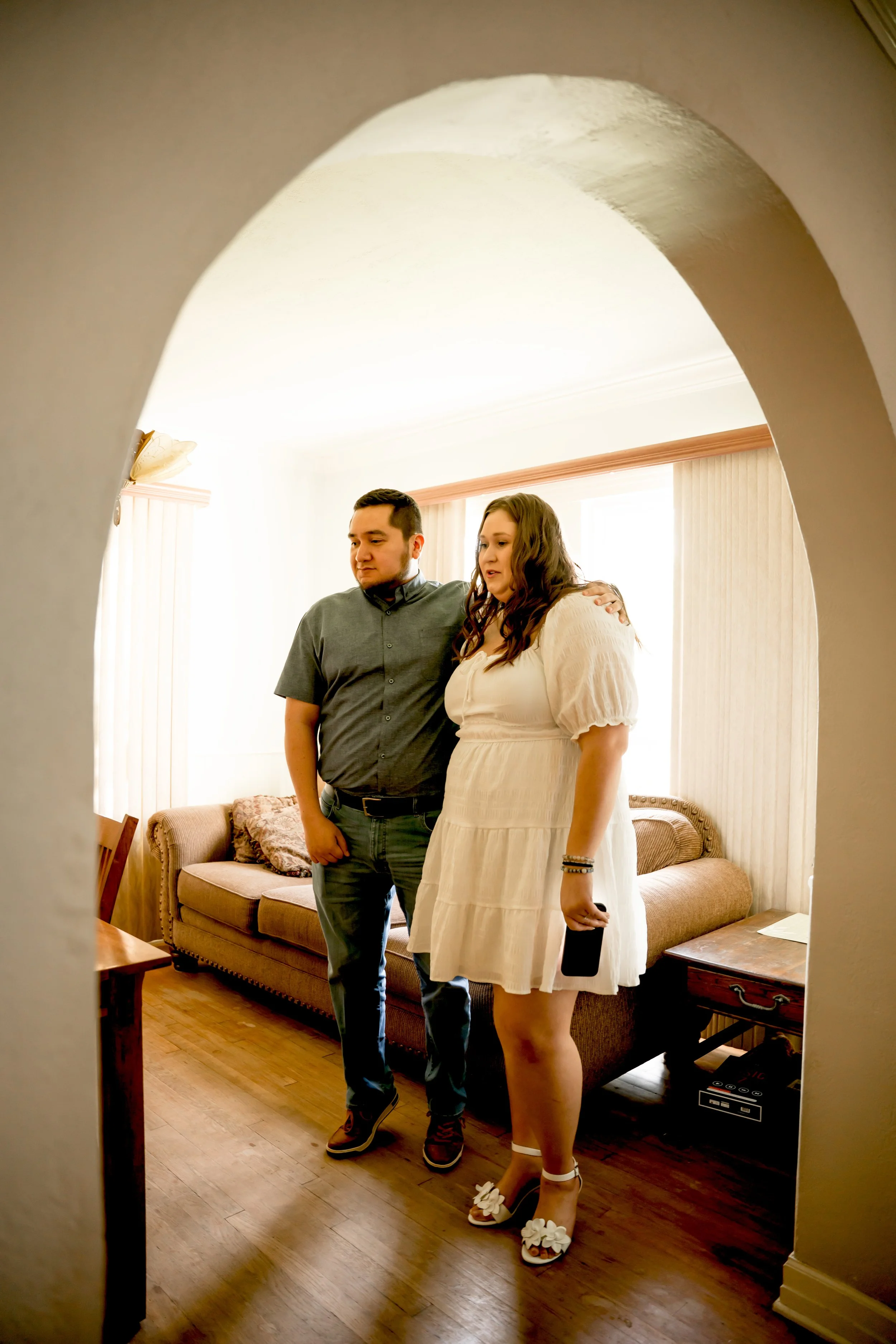 A man and a pregnant woman standing close together in a living room, with the man gently holding her shoulder, seen through an arched doorway.