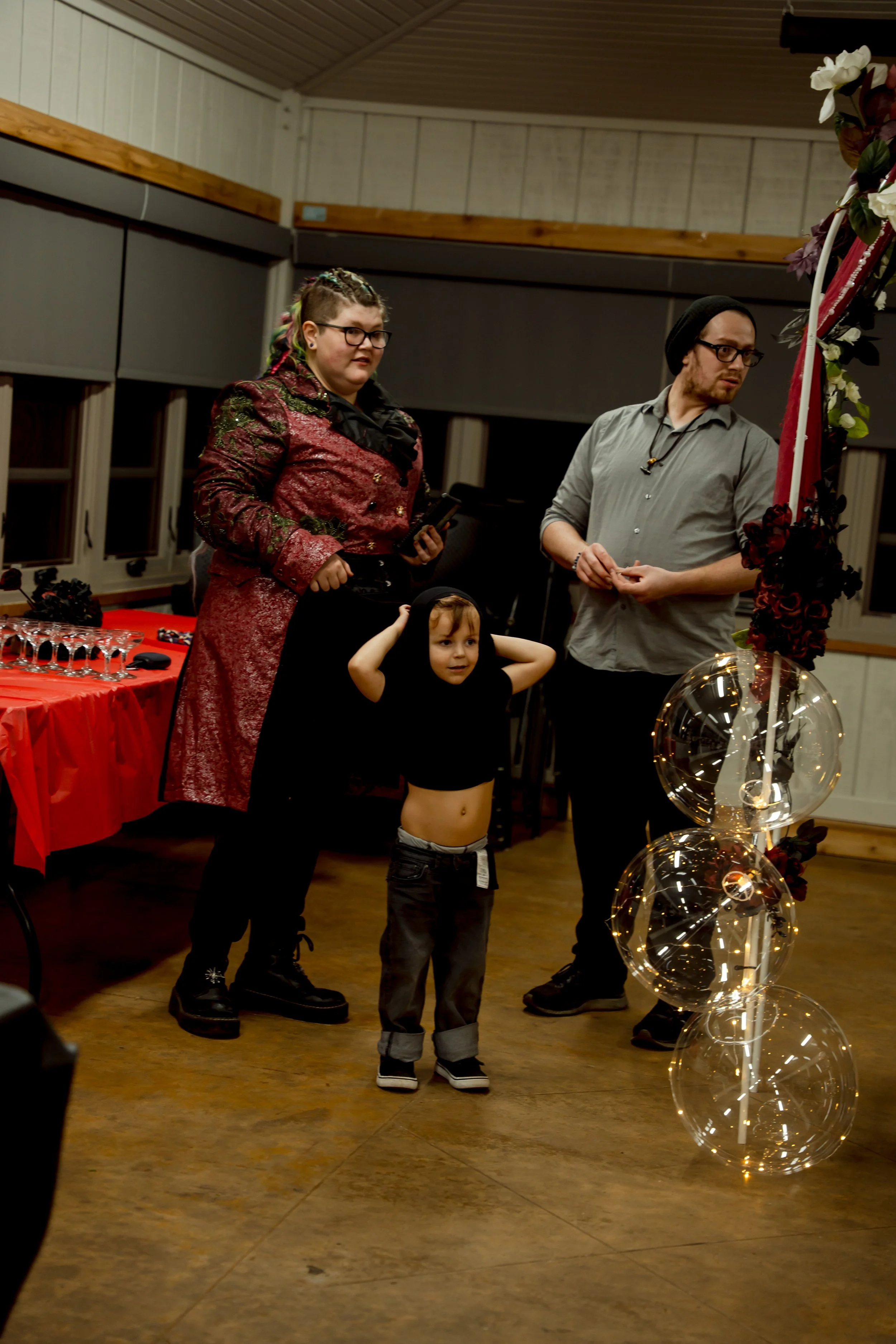A group of three people, two adults and a child, standing indoors near a decorative balloon arrangement with spherical transparent balloons and fairy lights. The woman on the left is dressed in a red and black patterned jacket, the man on the right i