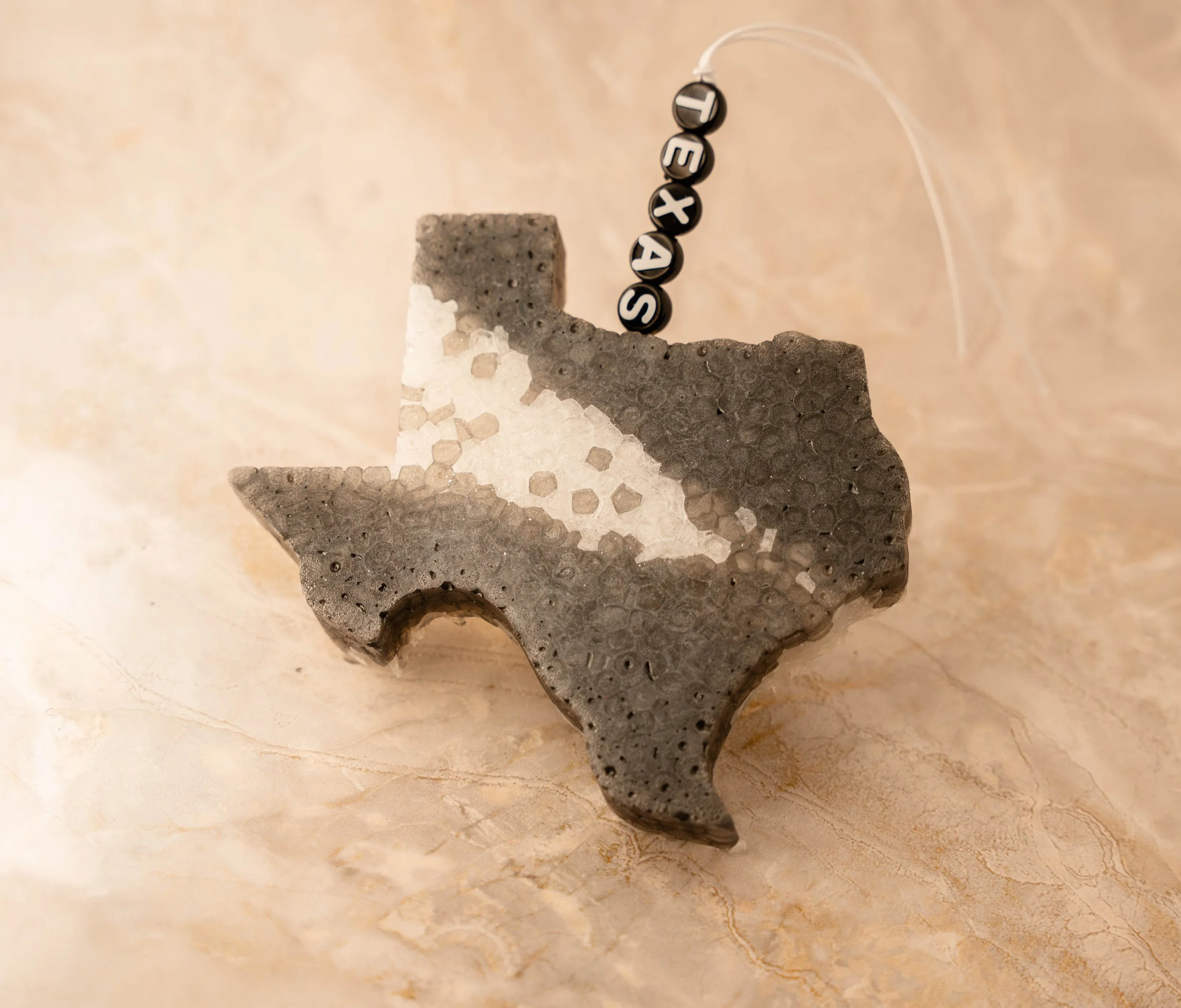 A decorative Texas-shaped object made from gray and white material, with a string of black beads spelling out 'TEXAS' hanging above it.