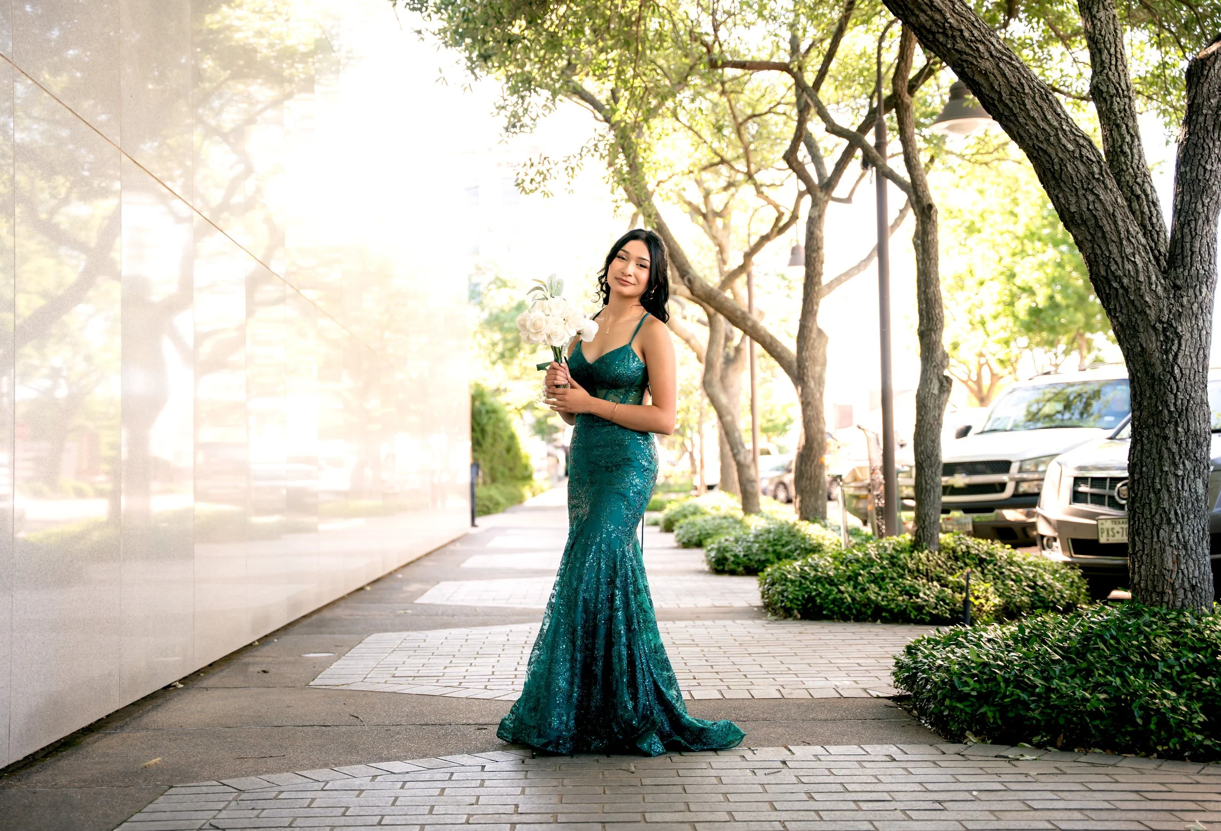 A young woman in a teal, sparkling floor-length dress holds a white bouquet on a city sidewalk with trees and parked cars in the background.