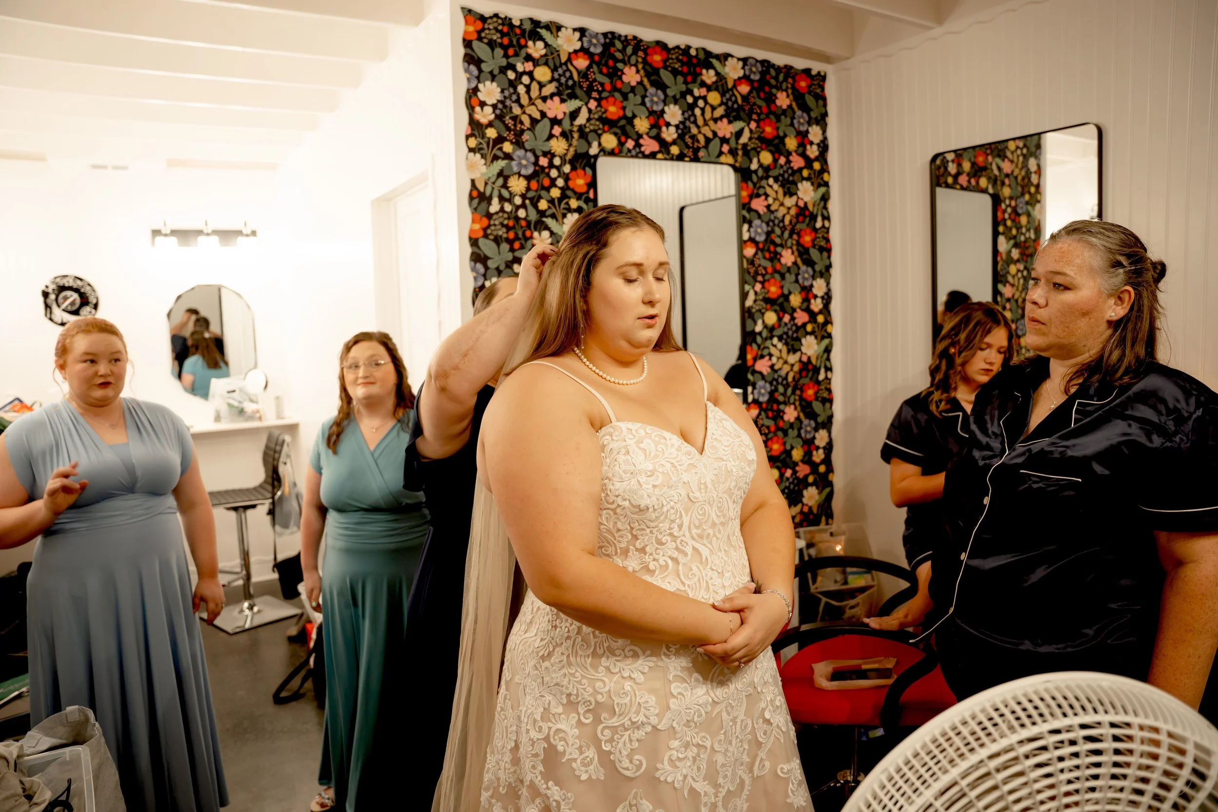 A bride in a white lace wedding dress with a pearl necklace is getting her hair fixed by a woman in black pajamas, while four other women in dresses watch in a room with floral wallpaper and mirrors.
