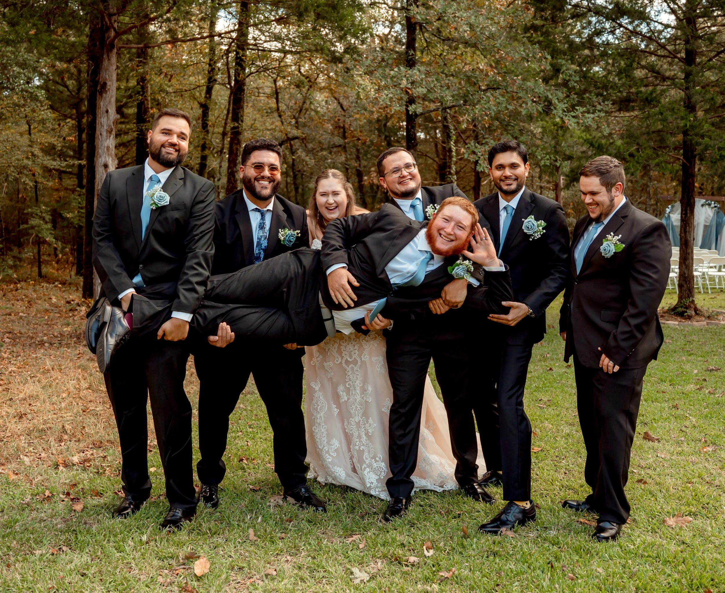 A group of eight people dressed in formal attire, with men in suits and the woman in a bridal gown, outdoors in a wooded area. They are smiling and laughing, and one man is being held horizontally by others, creating a playful scene.