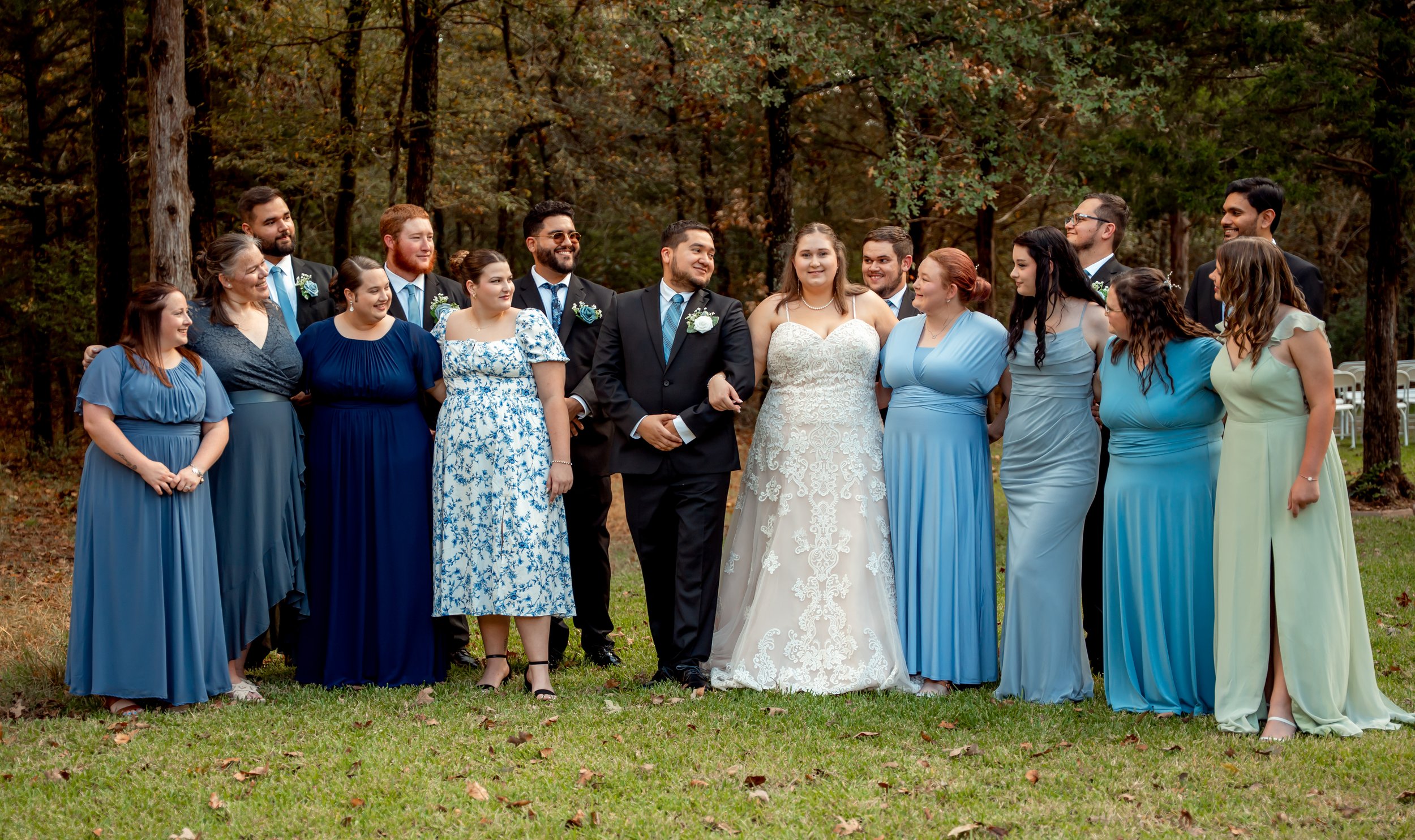 Group of bridal party and wedding couple standing outdoors in a wooded area, dressed in formal attire, smiling and looking at each other.