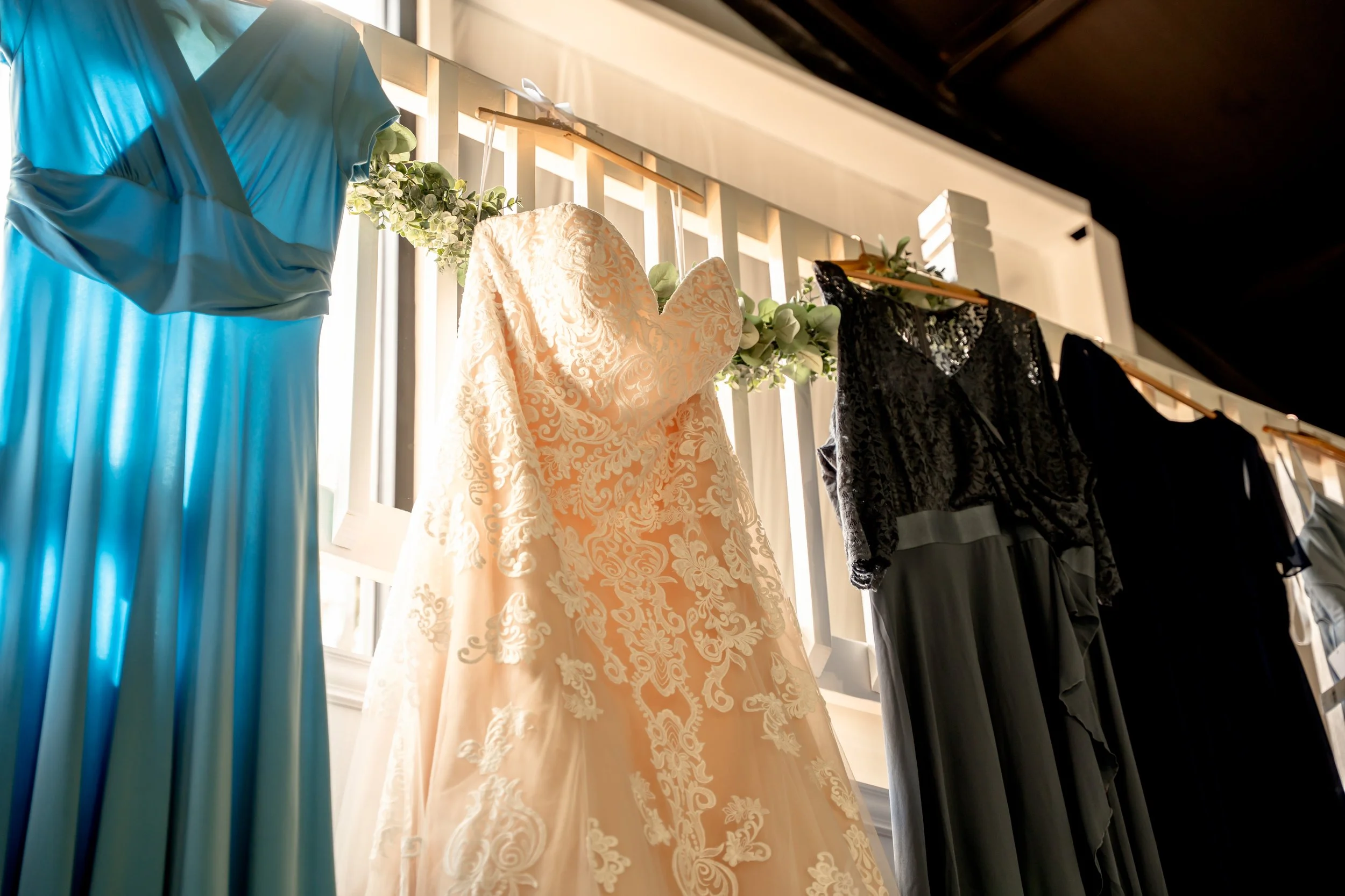 Various dresses hanging on a rack, including a peach wedding gown with lace details, a blue dress, a black dress with lace, and a dark-colored dress, displayed indoors with natural light 