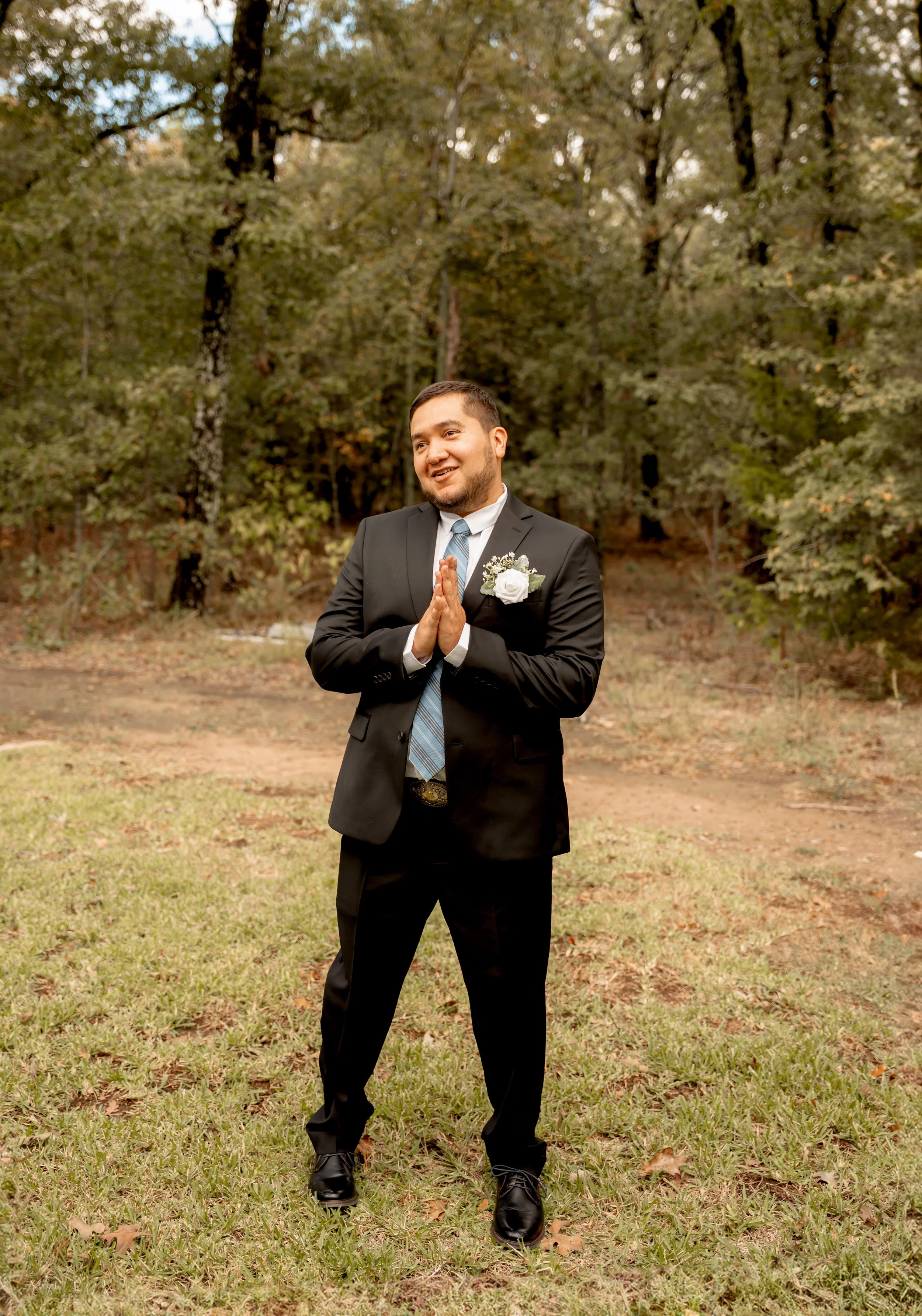 Man in a suit standing outdoors on grass, smiling with hands pressed together, surrounded by trees.