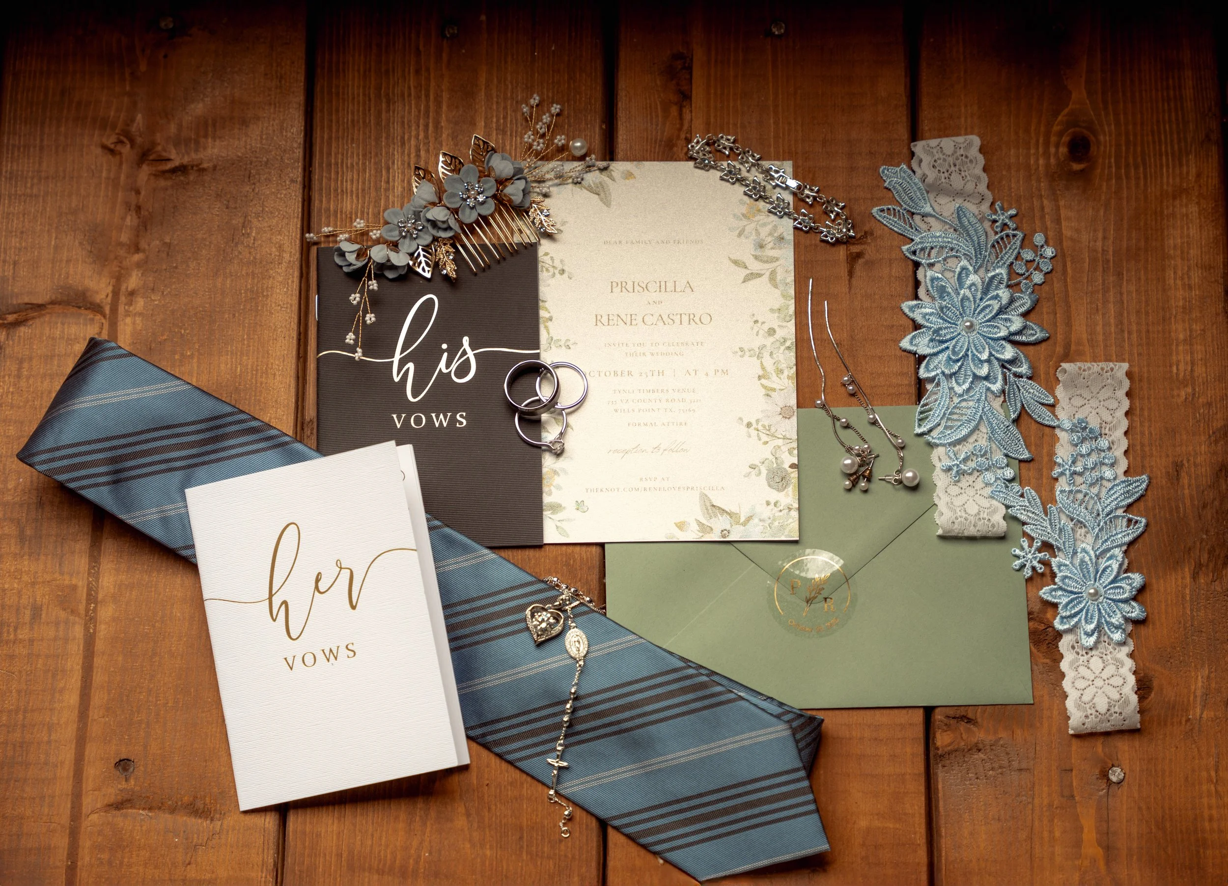 Wedding invitation suite and accessories arranged on a wooden surface, including invitations, vow book, jewelry, lace trim, and hair accessories.