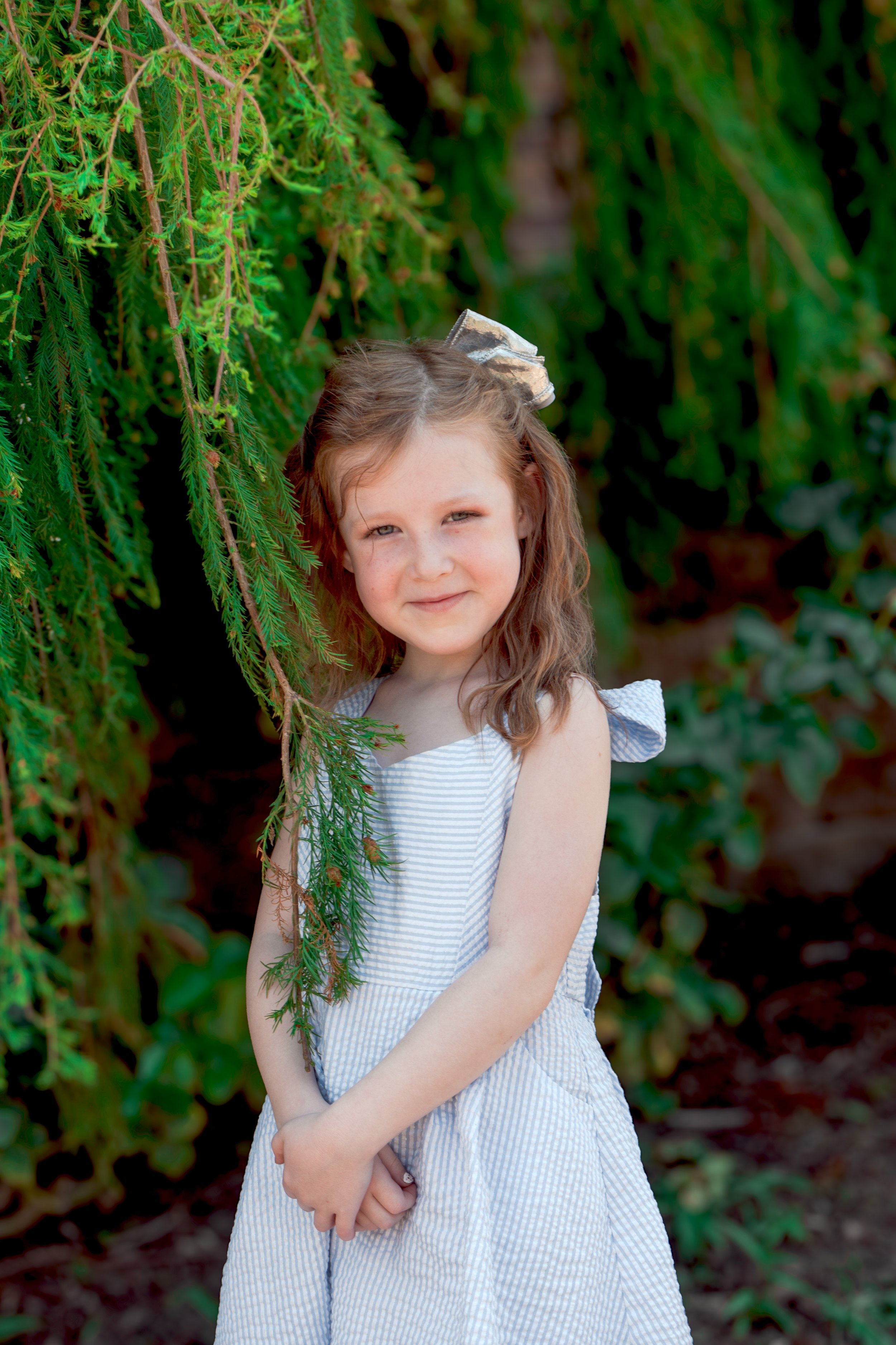 A young girl with brown hair and light skin, wearing a light blue and white striped dress, standing outdoors amidst green foliage.