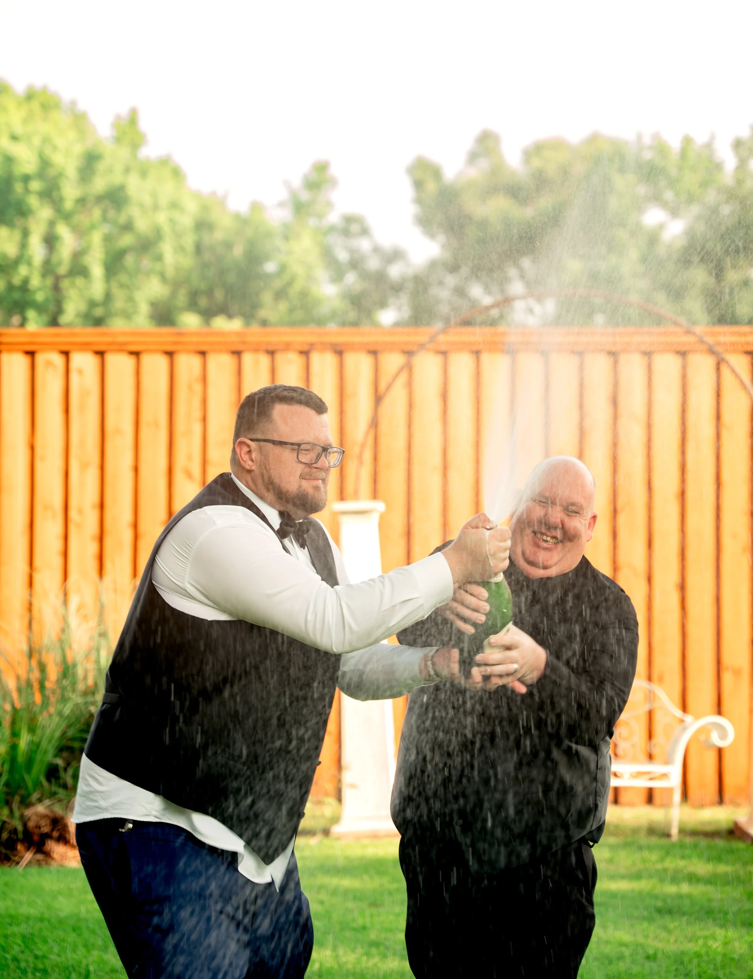 Two men in tuxedos celebrating with a champagne bottle in a backyard with a wooden fence, benches, and greenery, as champagne sprays out of the bottle.