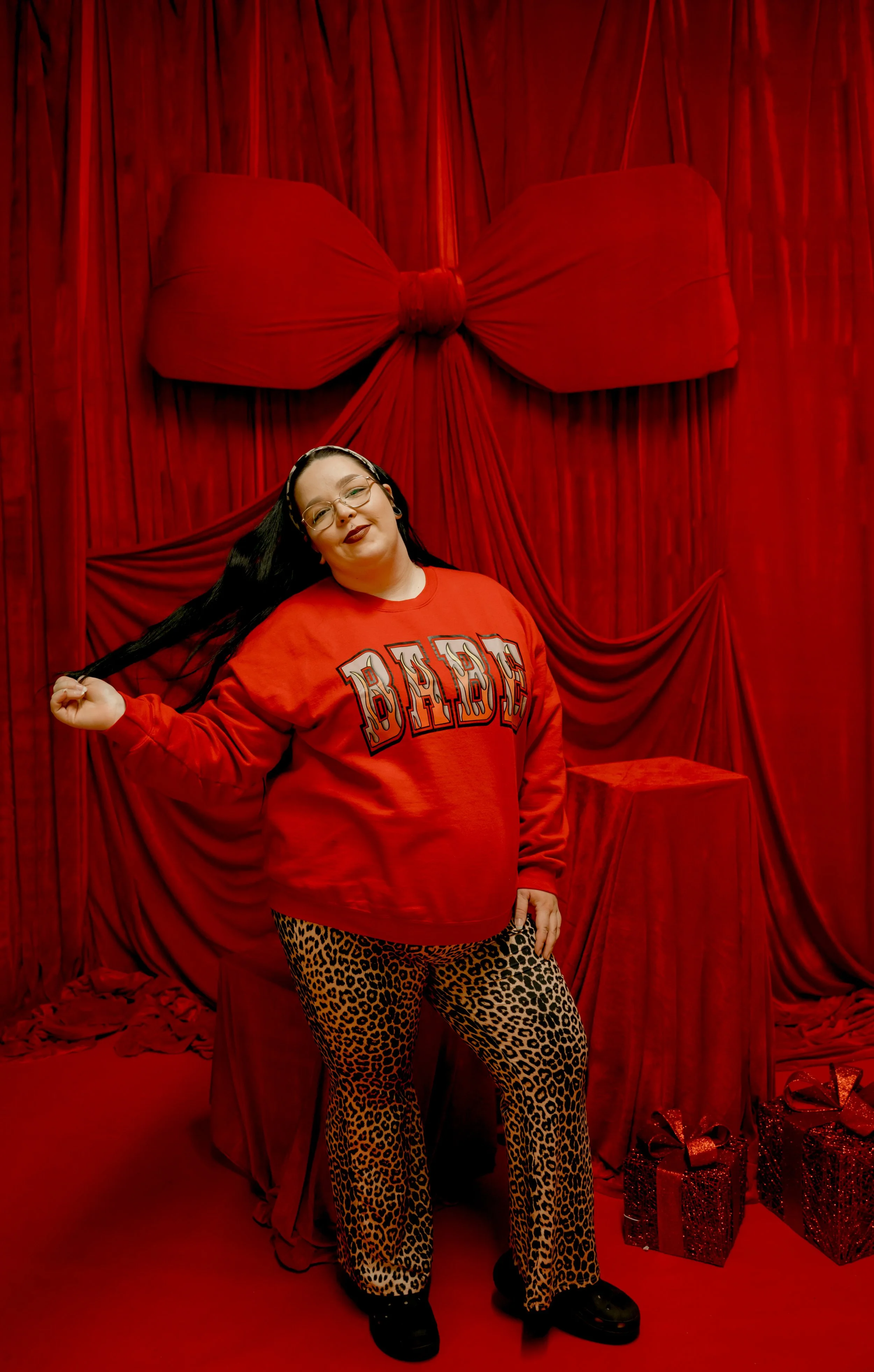Woman standing in front of red velvet drapes with wrapped presents at her feet, wearing leopard print pants and a red sweatshirt.