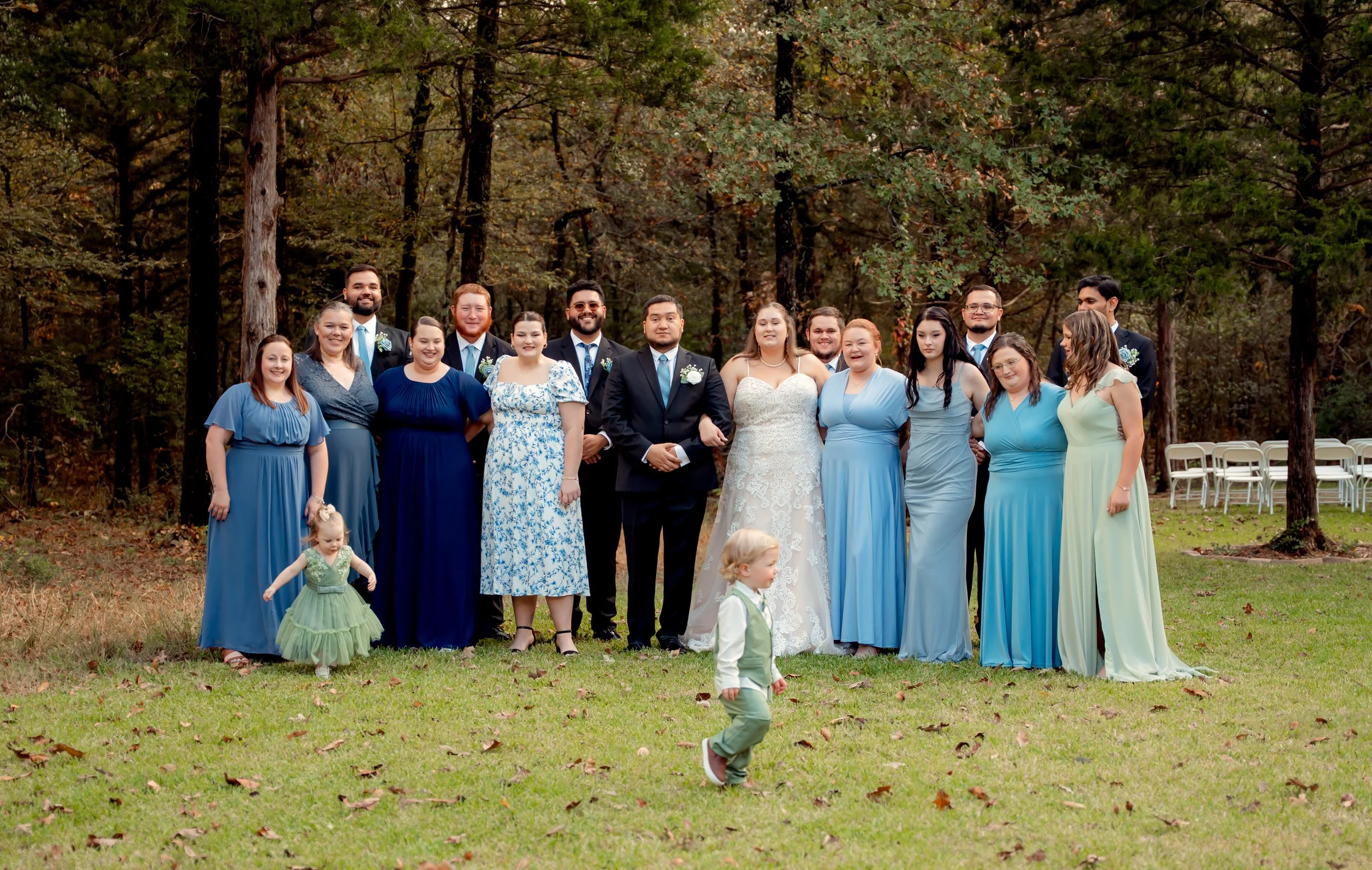 A wedding party posing outdoors in a wooded area with trees. The group includes a bride and groom, bridesmaids, groomsmen, and two young children. The women are wearing various shades of blue and pastel dresses, while the men are in suits with ties. 