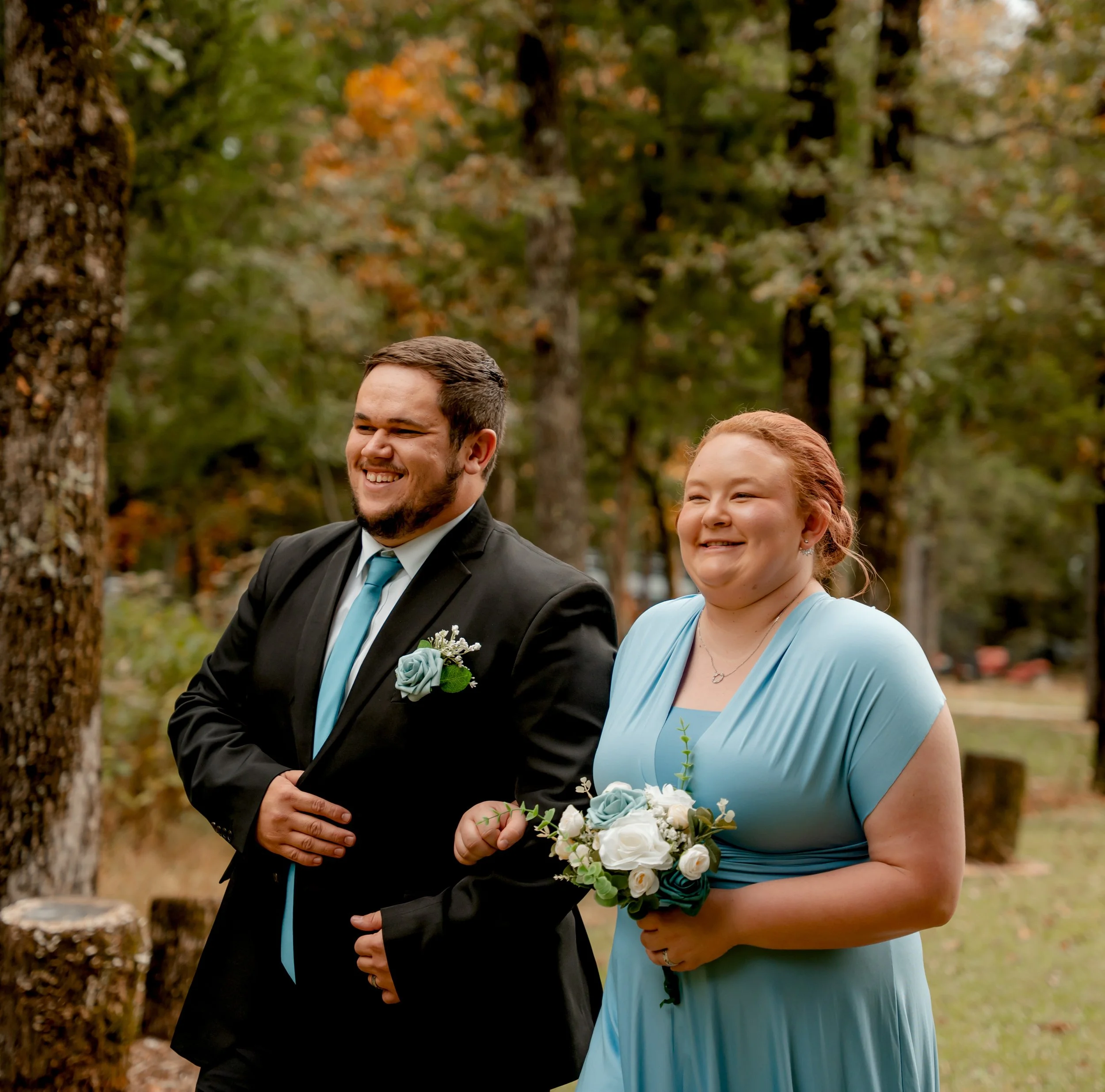 A smiling man in a black suit and blue tie walking arm-in-arm with a smiling woman in a light blue dress holding a bouquet of white and blue roses, outdoors with trees in the background.