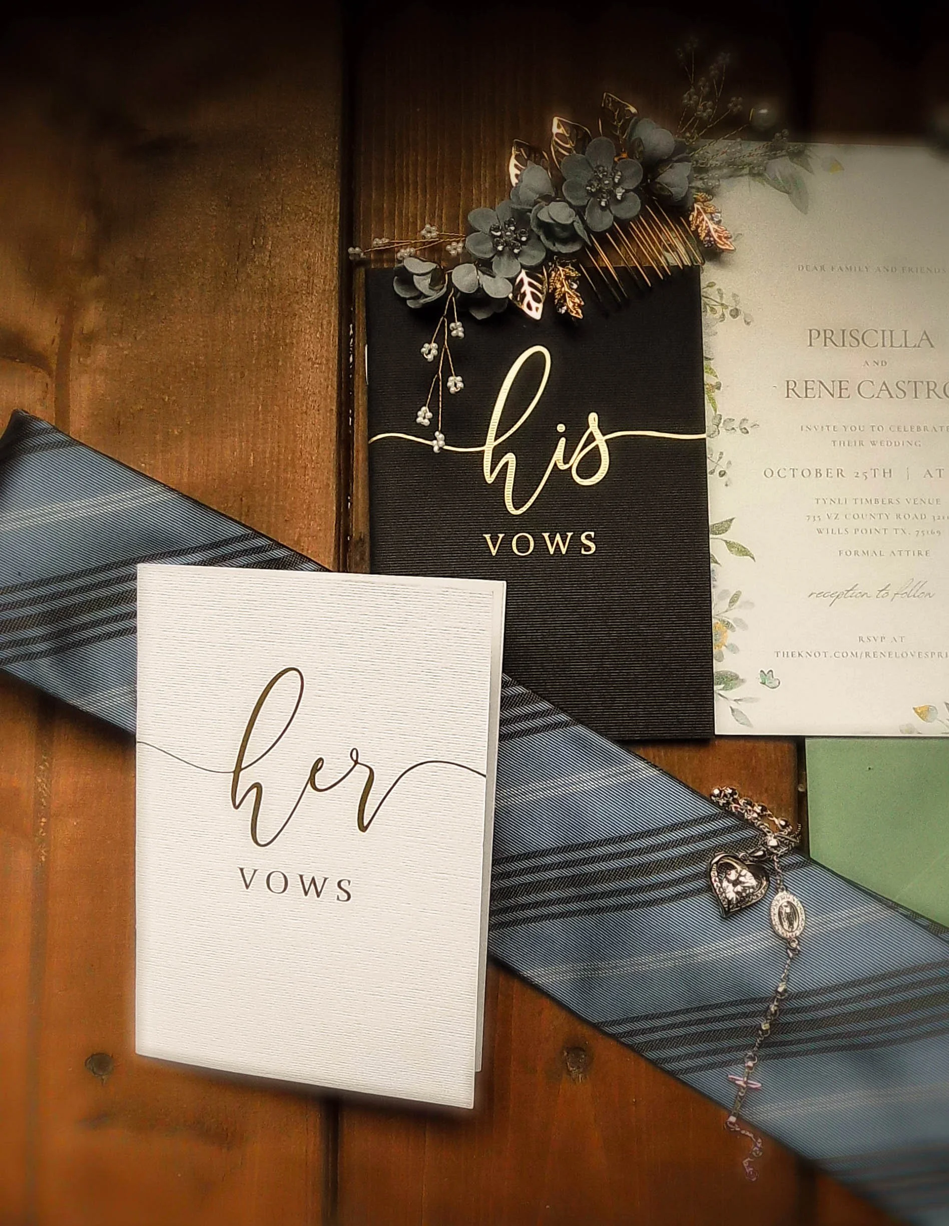 Wedding vows booklets labeled "her" and "his" on a wooden table, along with a floral hair comb, a rosary or bracelet, and wedding invitation card with floral design.