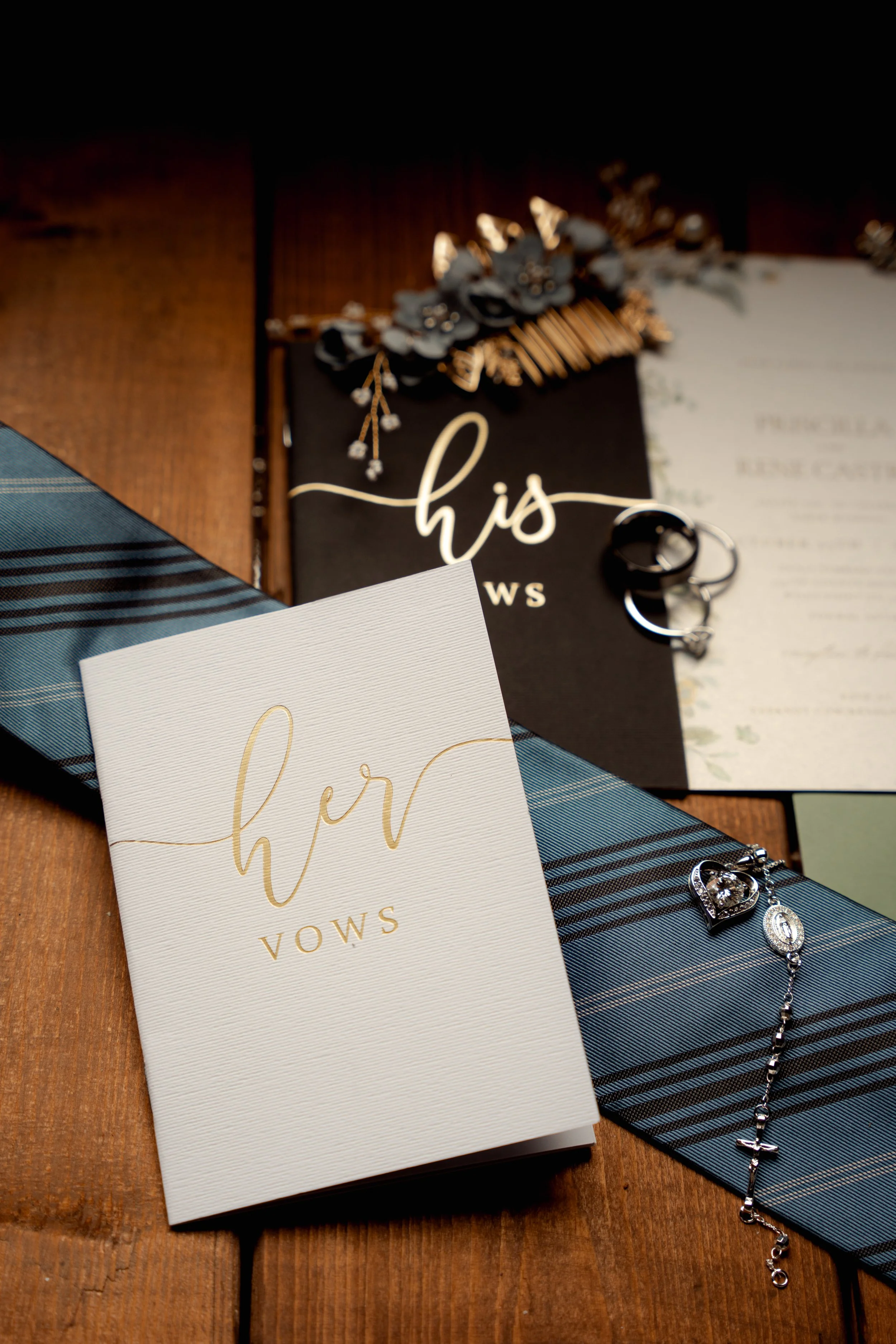 Wedding vow booklets, wedding rings, a blue patterned ribbon, and jewelry arranged on a wooden surface.