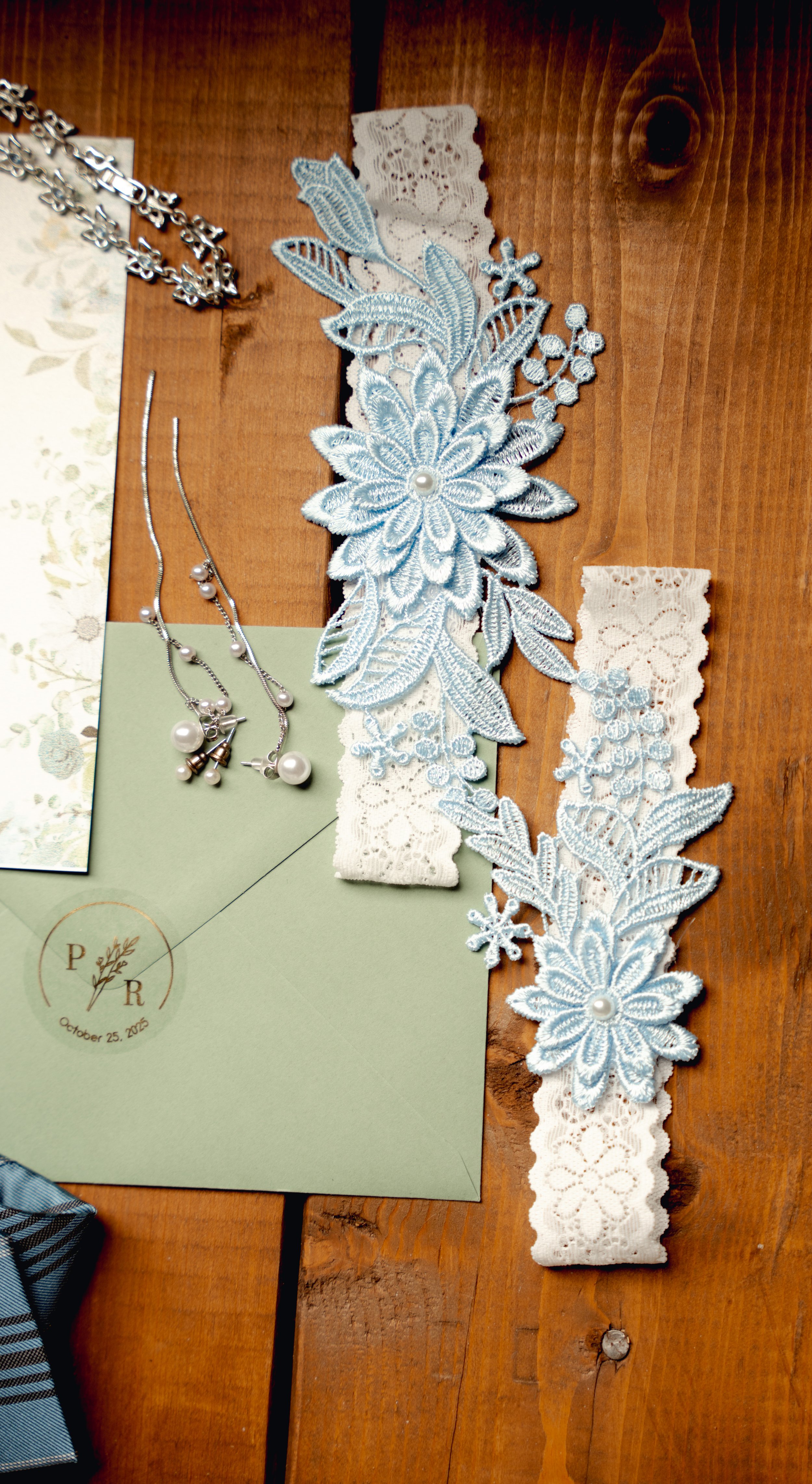 Light blue floral lace appliqué with pearl accents, paired with white lace headbands, pearl earrings, and a green wedding invitation with a monogram and date.