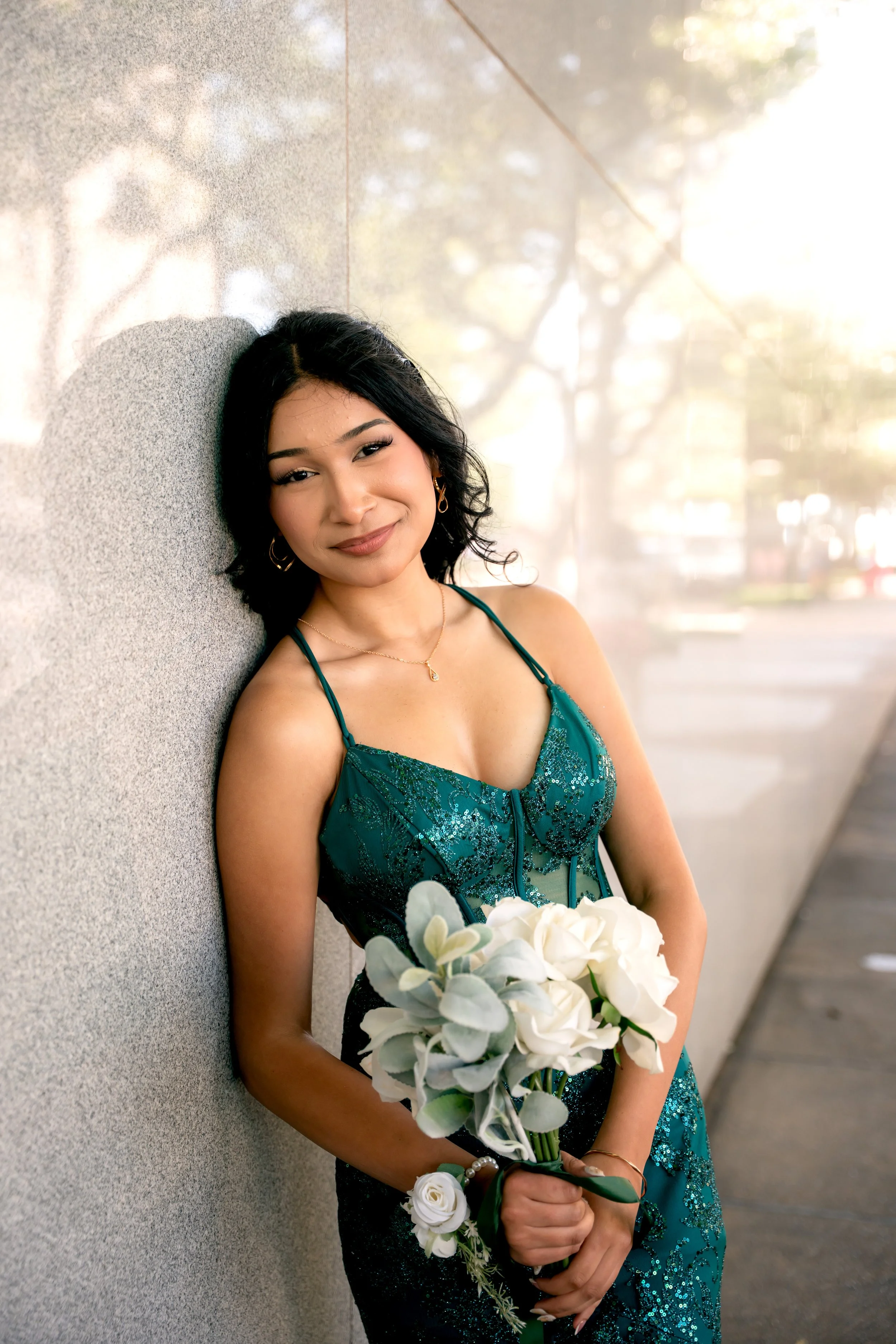 Young woman in a teal dress holding a bouquet of white flowers, leaning against a textured wall outside, smiling at the camera.