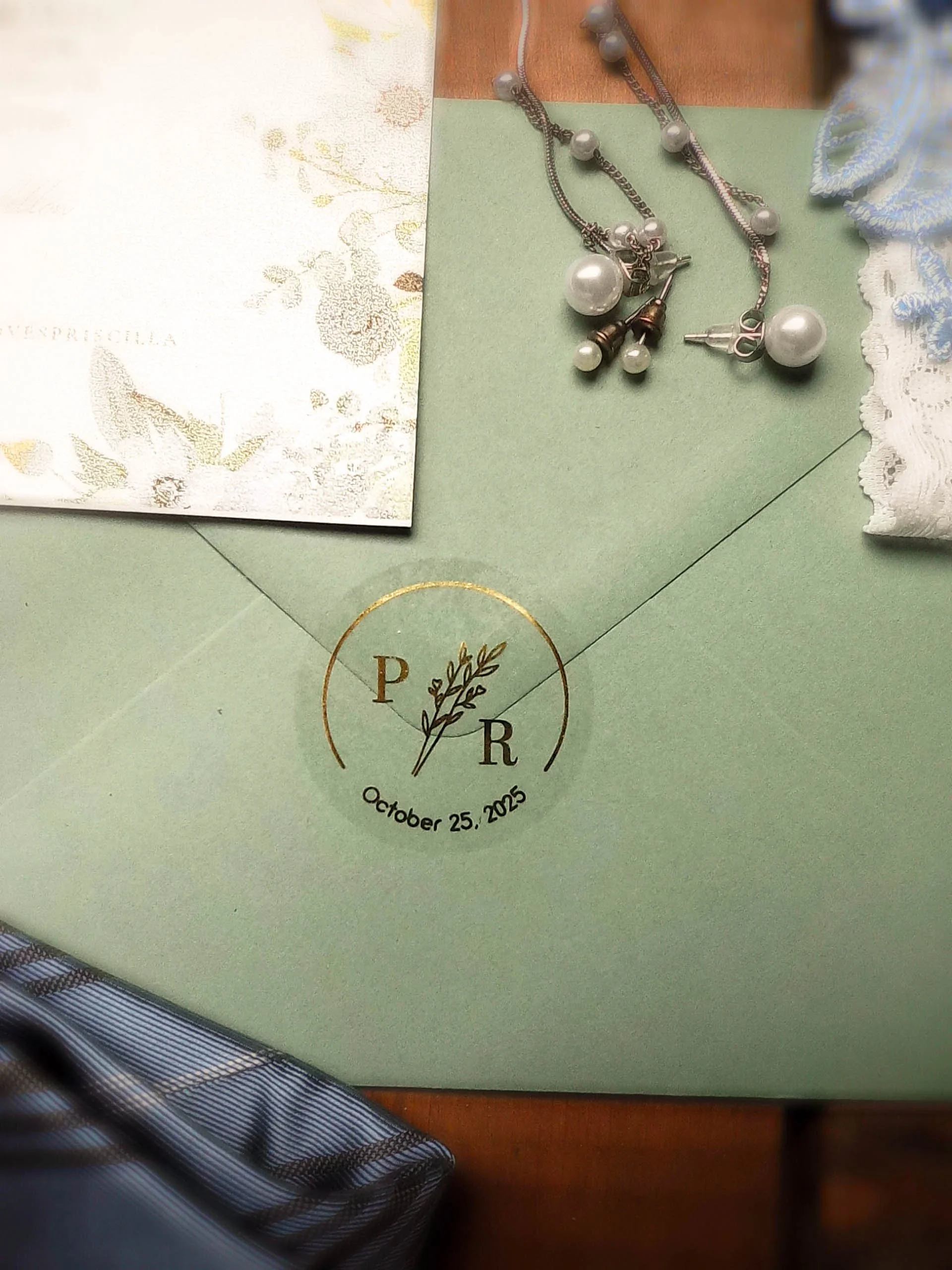 Wedding invitation with jewelry, including pearl earrings and necklace, on a green cloth with a white lace cloth nearby. The invitation has the initials P and R, a floral design, and the date October 25, 2025.