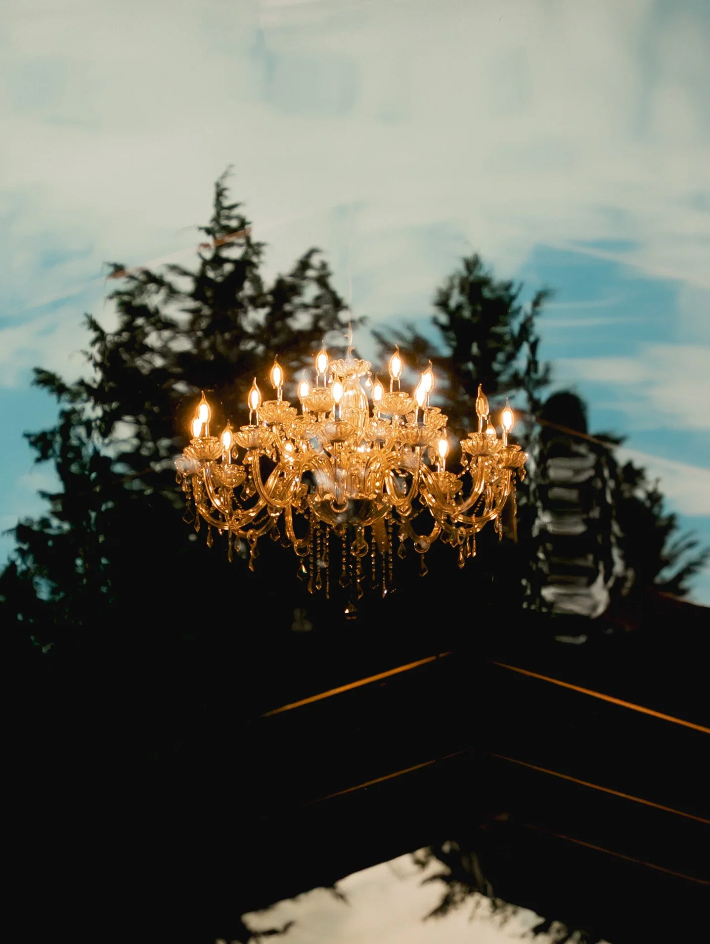A sparkling chandelier hanging outdoors with a background of trees and a partly cloudy sky at dusk.