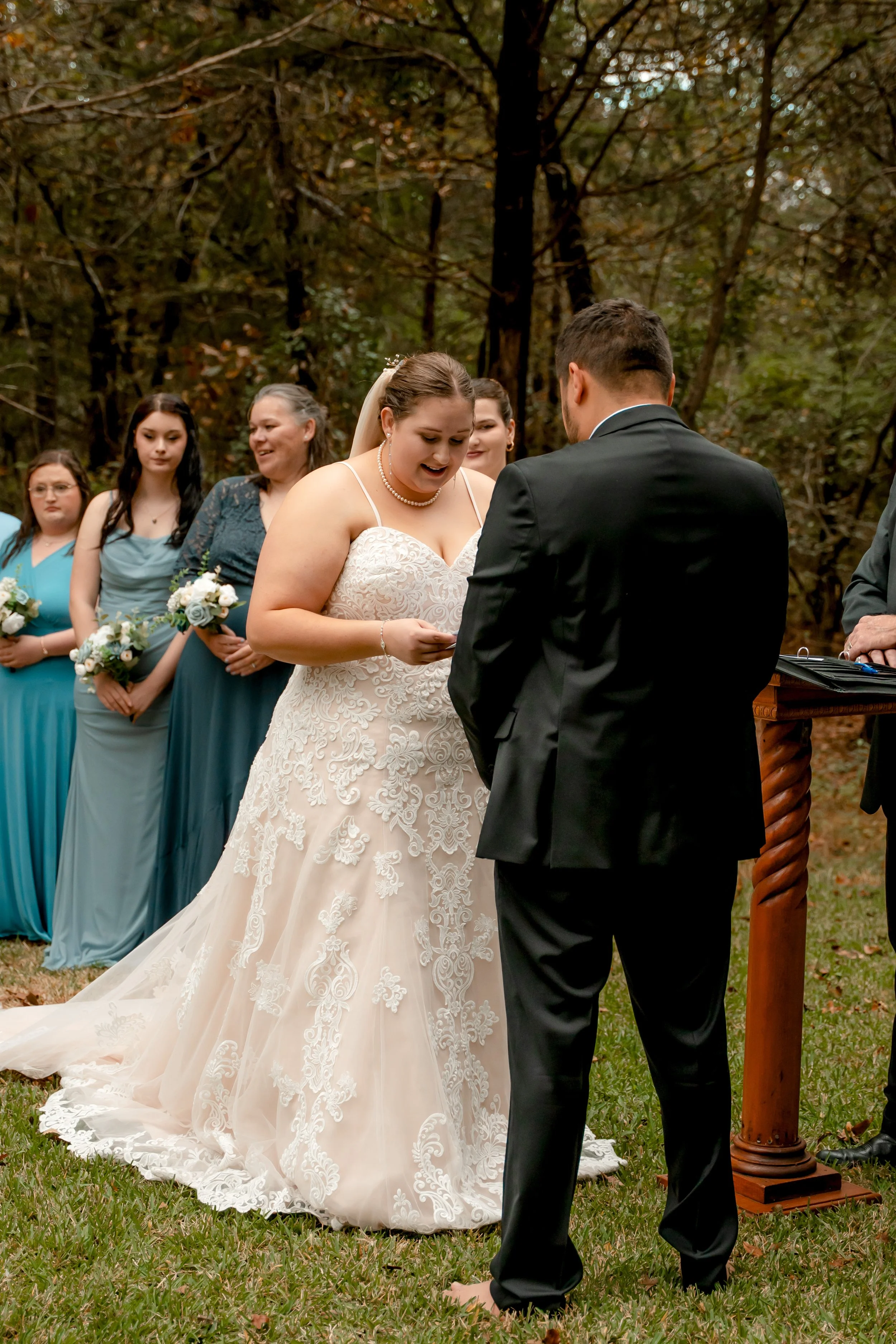 Woman in wedding dress exchanging vows with man in black tuxedo during outdoor wedding ceremony in wooded area, surrounded by bridesmaids in blue dresses.