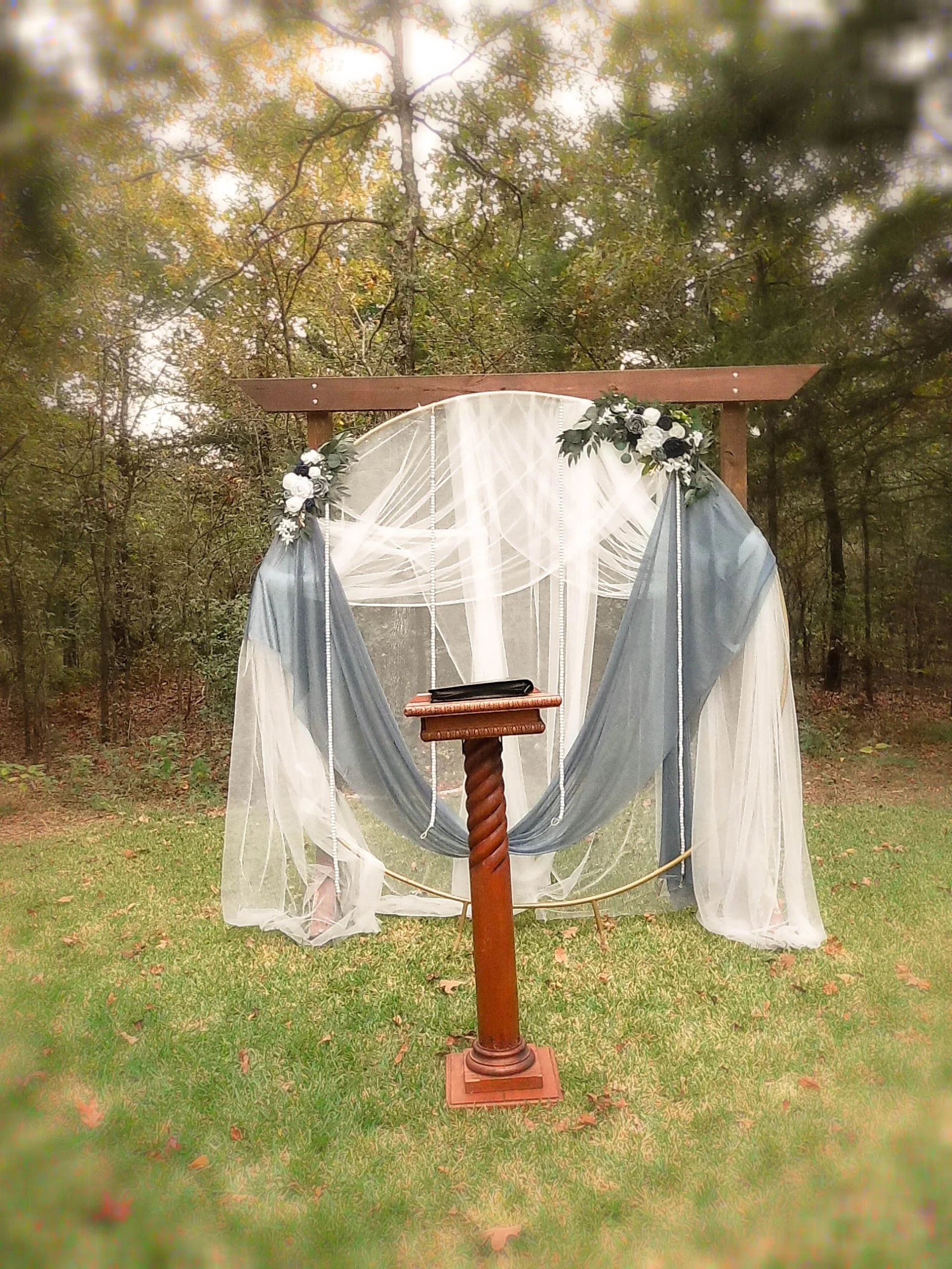Outdoor wedding altar with floral arrangements, draped white and blue fabric, and a small table in front on a grassy area surrounded by trees.