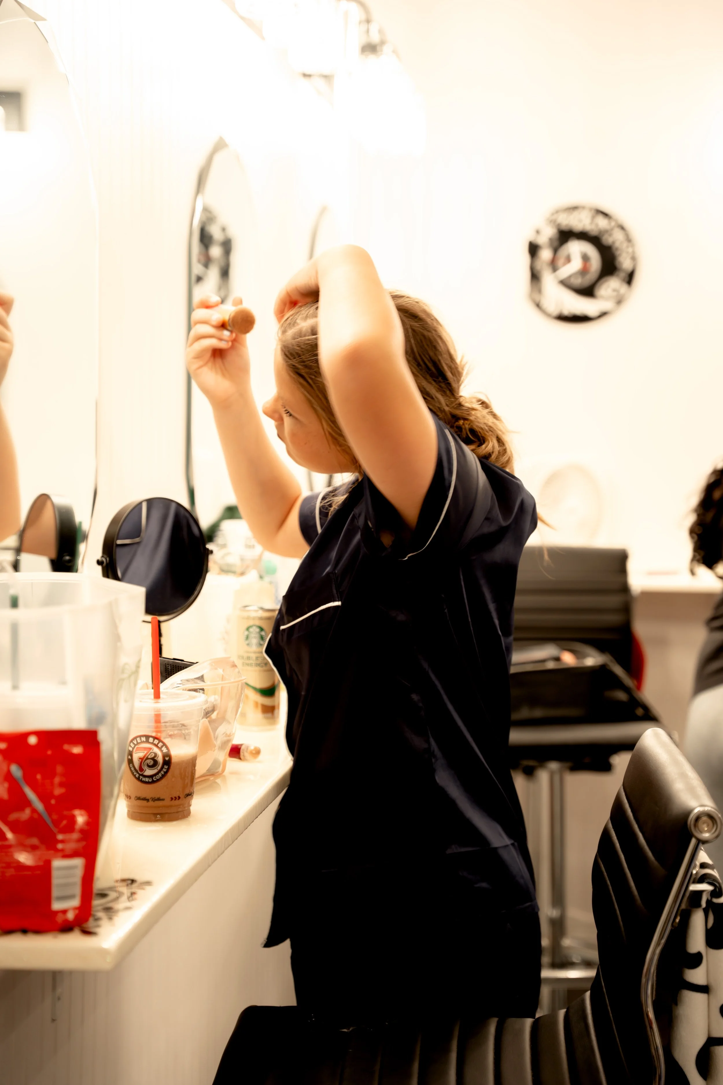 Woman with curly hair styling her hair at a bathroom counter, surrounded by makeup and coffee cups.