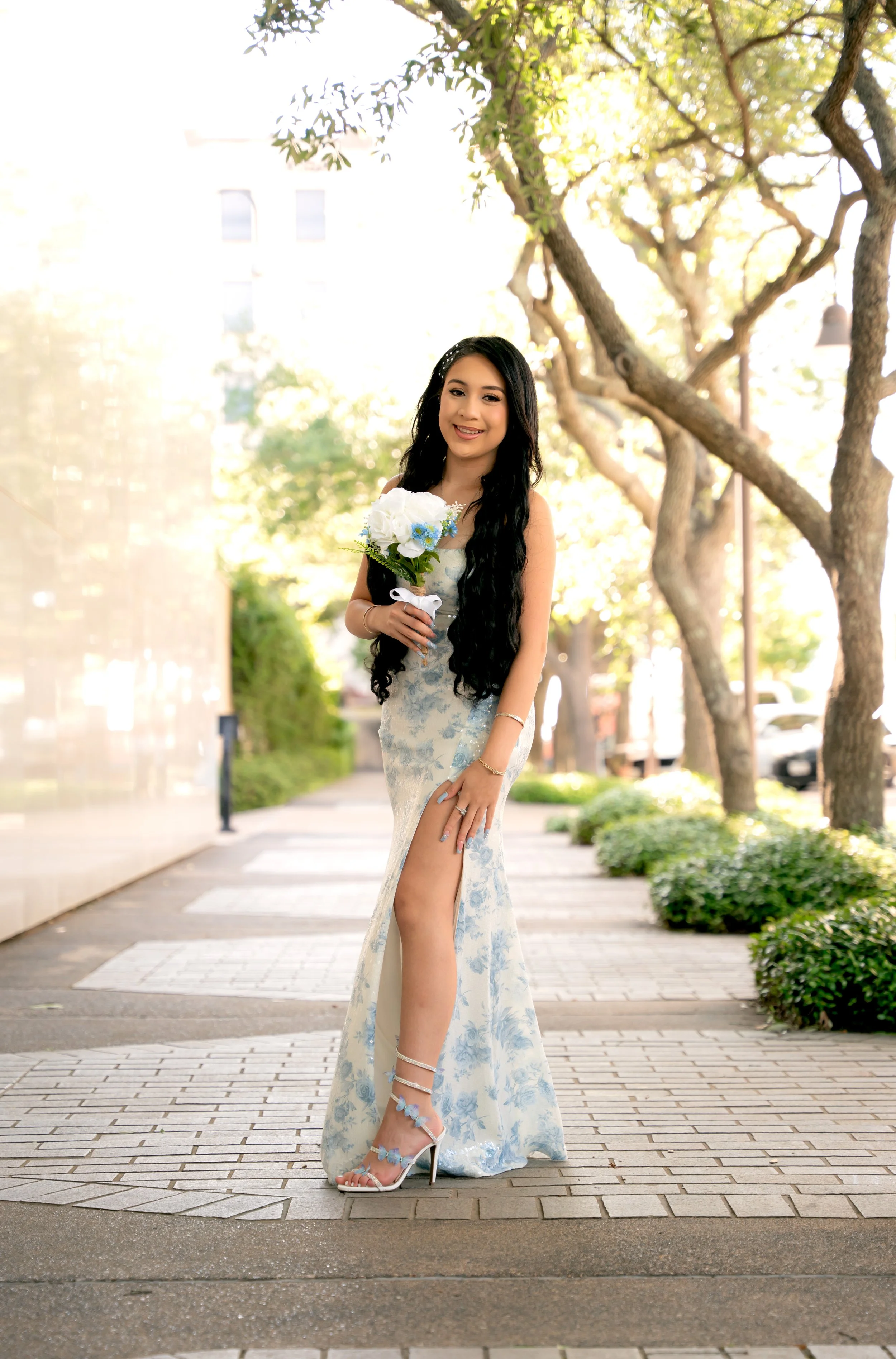 A woman in a white and blue floral gown holding a bouquet of white and blue flowers, standing on a city sidewalk with trees and buildings in the background, smiling at the camera.
