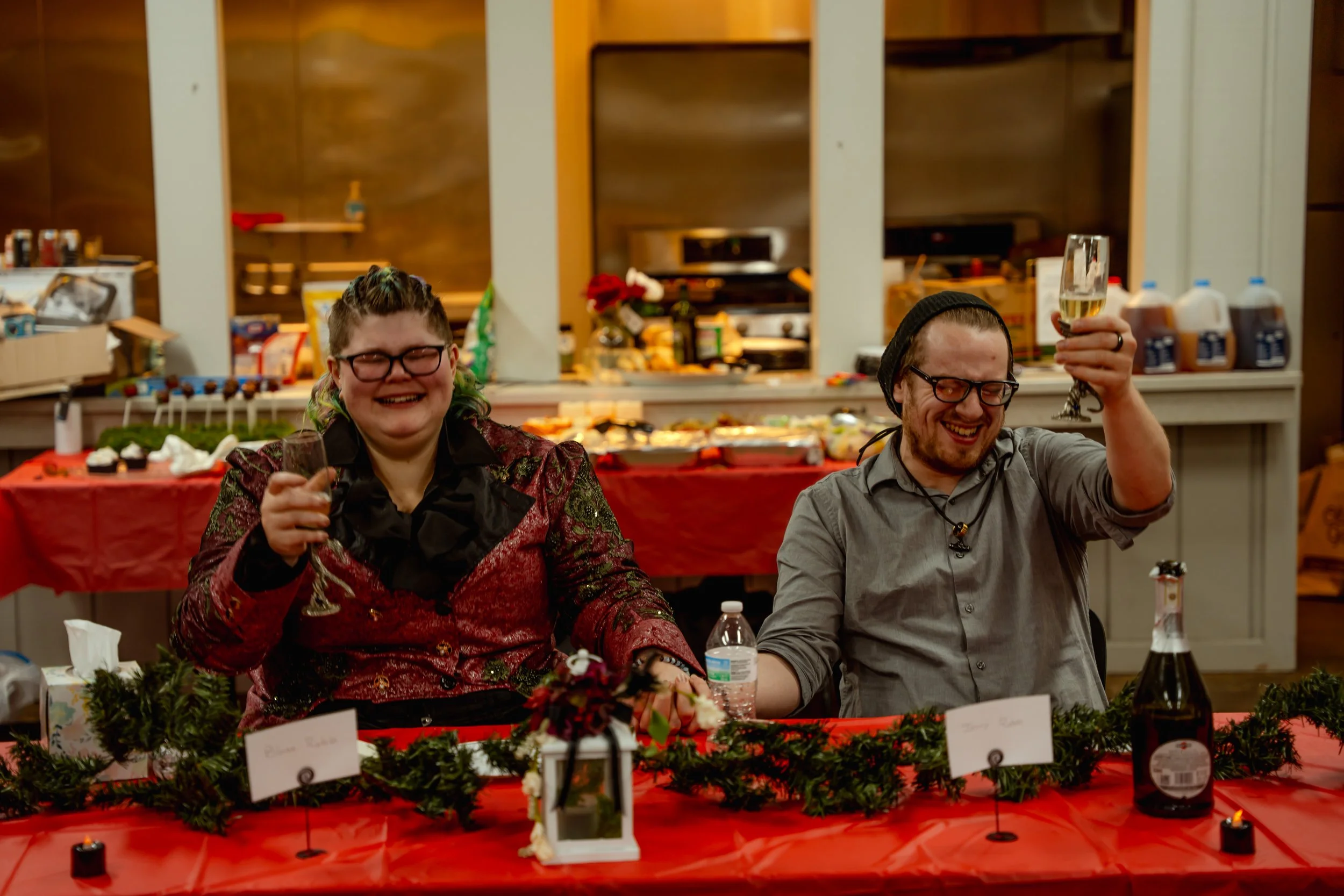 Two people sitting at a decorated table celebrating, holding drinks and smiling. Background shows a kitchen with food and drinks on a counter.