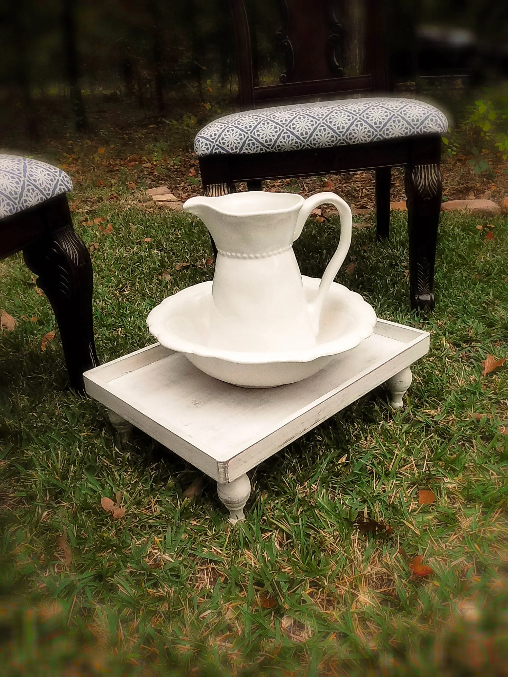 A white pitcher and bowl set placed on a small white wooden table outdoors, with a background of green grass and outdoor chairs.