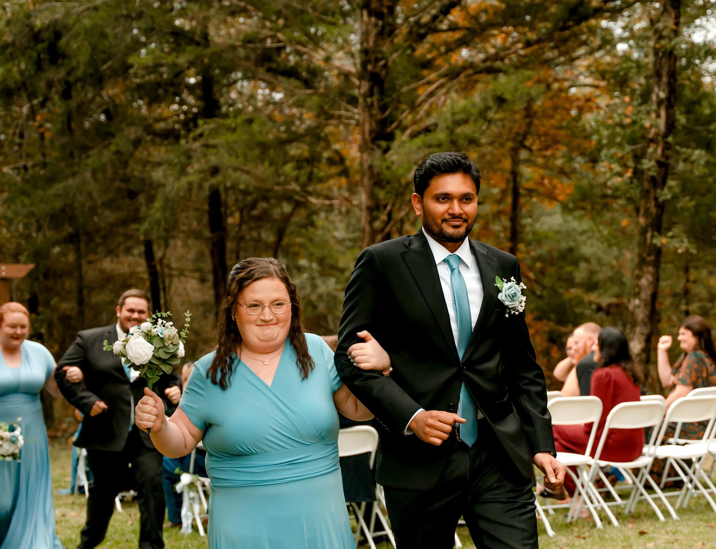 A wedding procession walking down an outdoor aisle in a wooded area during autumn. The bride, wearing glasses and a light blue dress, holds a bouquet and links arms with the groom, who is dressed in a black suit with a light blue tie and boutonniere.