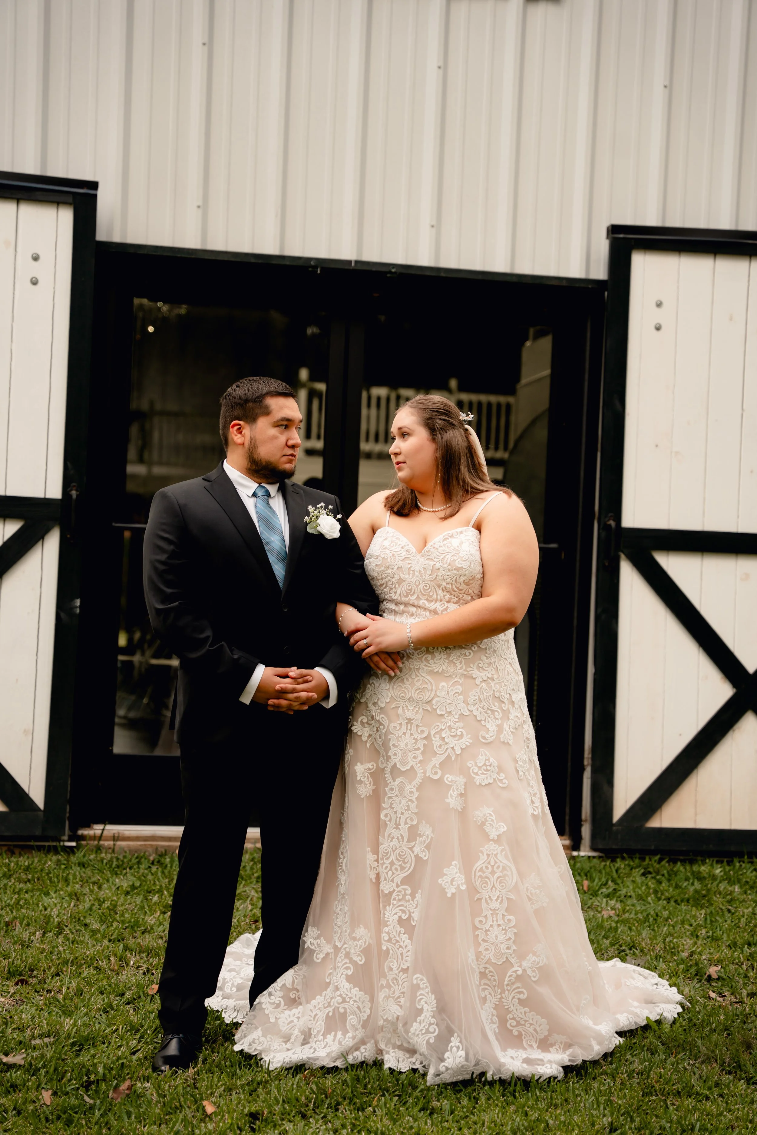 A bride and groom standing in front of a barn, looking at each other. The bride is wearing a lace wedding gown and the groom is in a black suit with a blue tie.