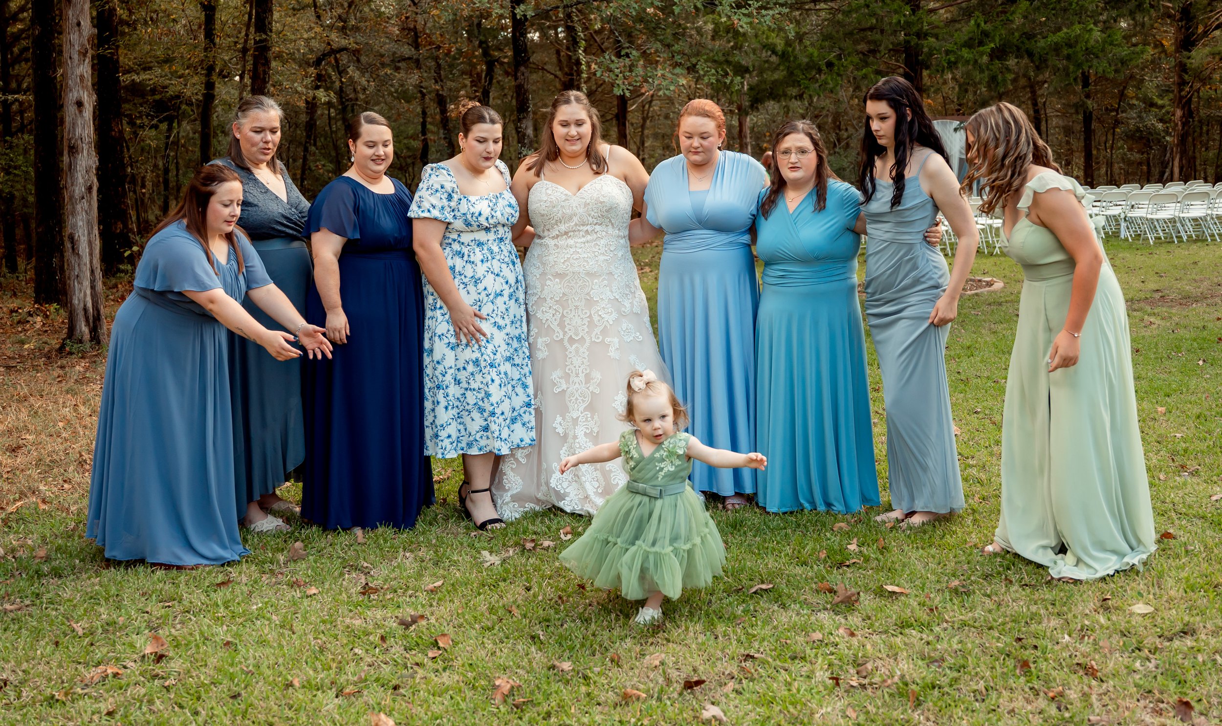 A group of women in various shades of blue dresses standing in a semi-circle outdoors, with a young girl in a green dress dancing in front of them, in a wooded area with chairs in the background.